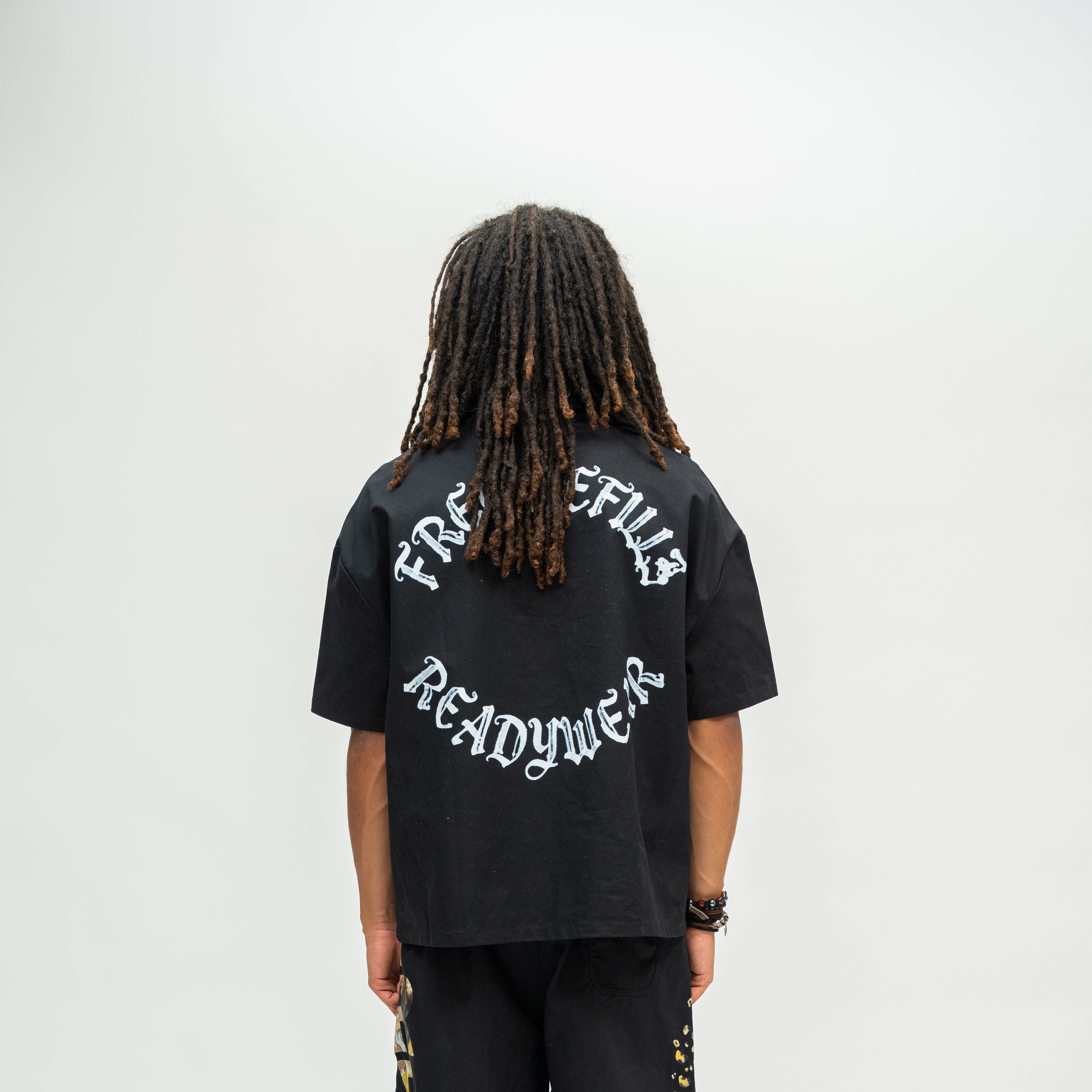 A person viewed from behind wearing a black oversized T-shirt with white Gothic-style text reading 'FREE REFILLS READYWEAR' across the back, paired with black pants, against a plain light gray studio background.