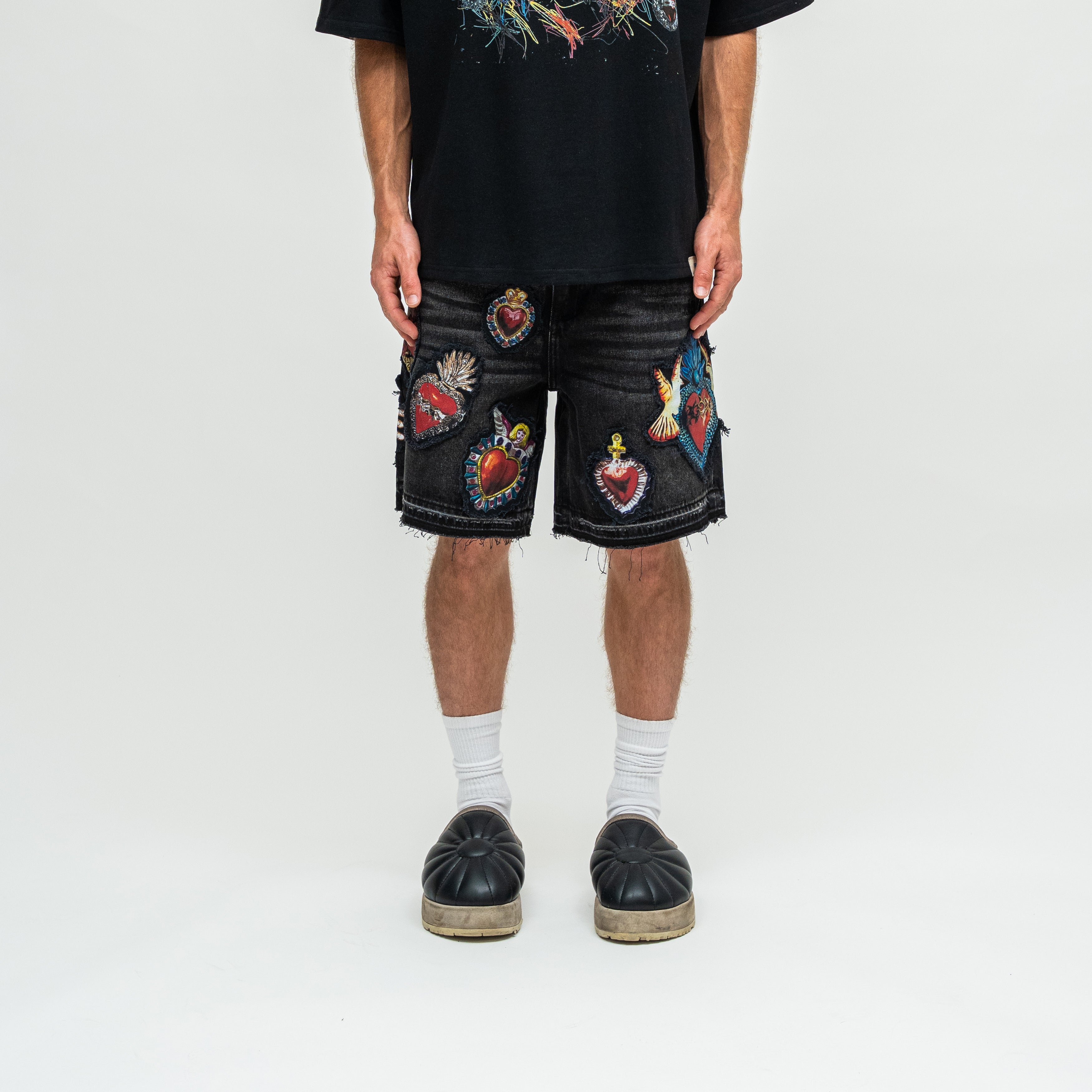 A person wearing black denim shorts covered with colorful embroidered patches, a black graphic T-shirt, white socks, and black espadrille-style shoes, photographed against a plain light gray studio background.
