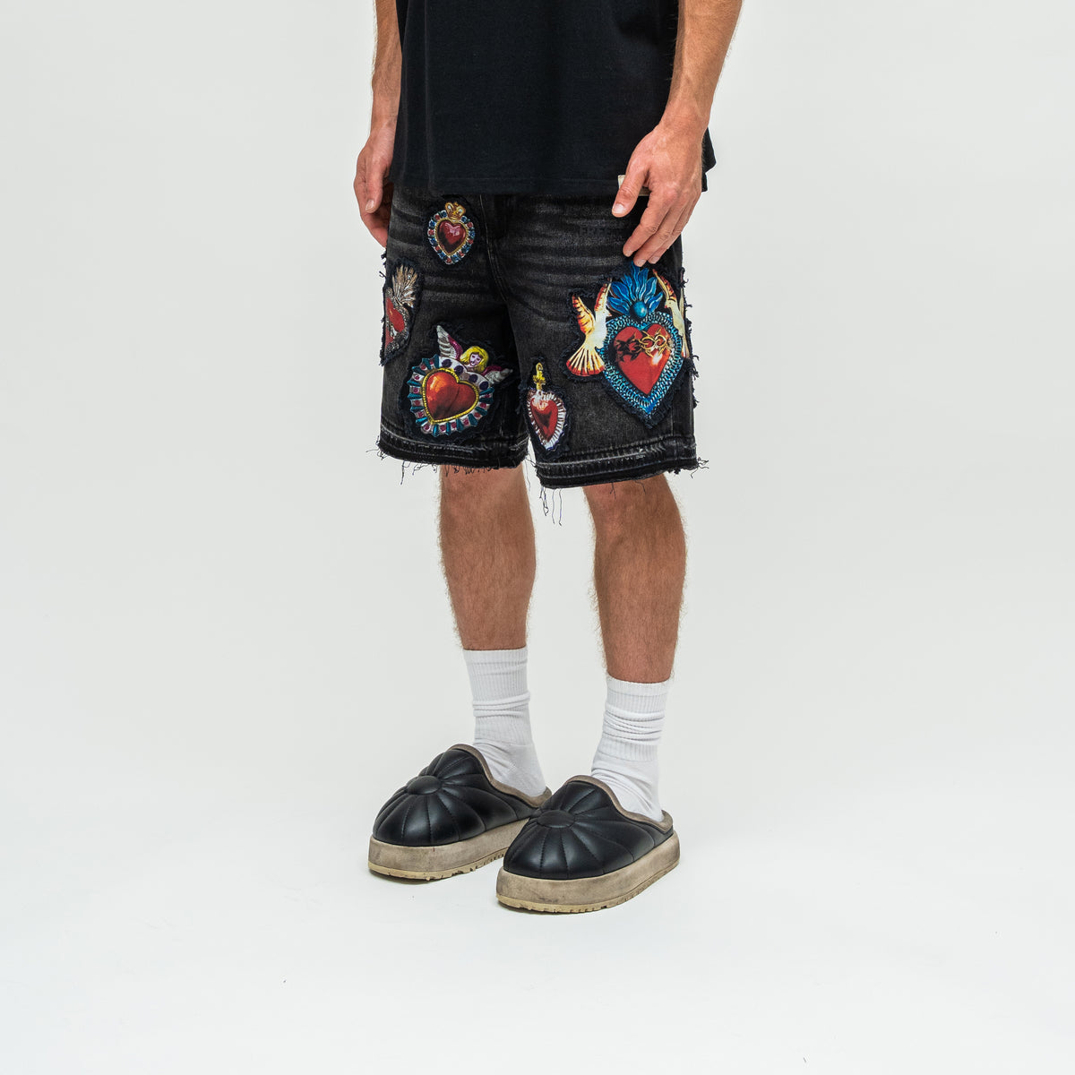 A person wearing black frayed denim shorts with colorful embroidered Sacred Heart patches, paired with white socks and black slip-on shoes, photographed against a light gray studio background.