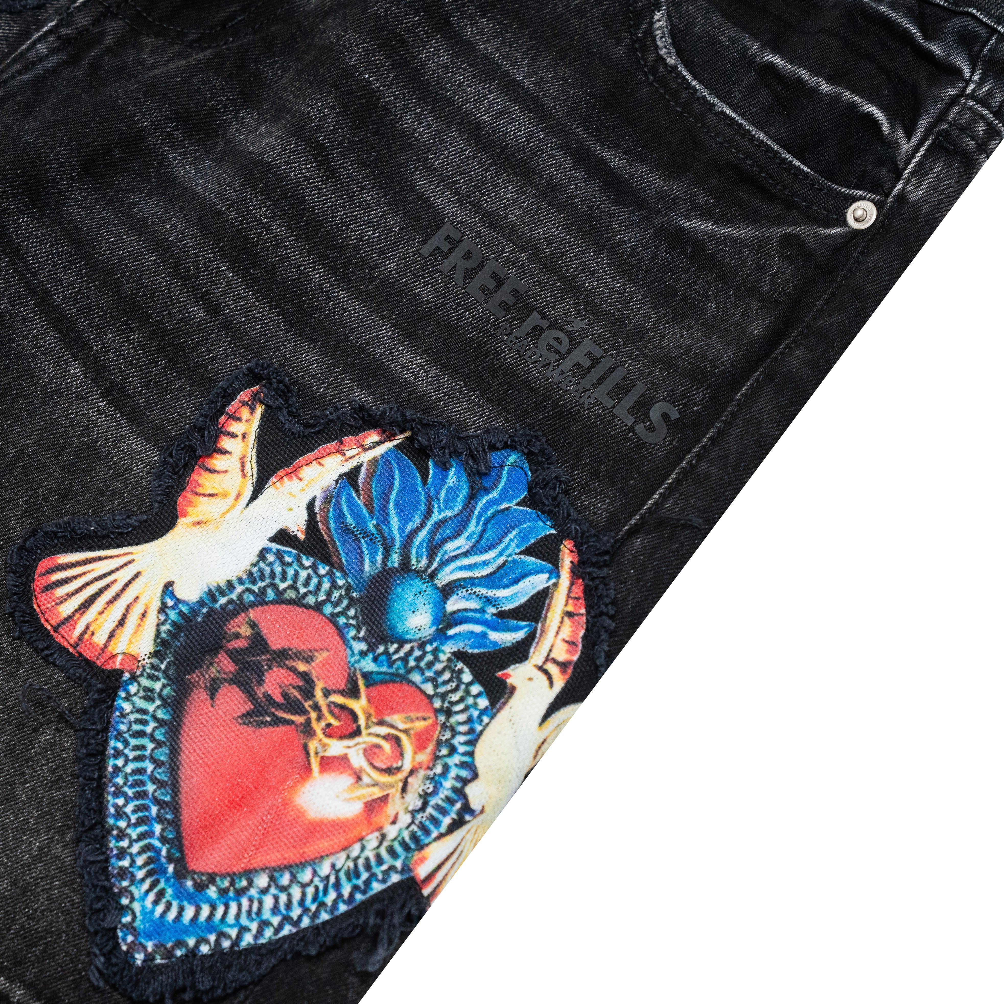 Close-up of black distressed denim jeans featuring a large embroidered patch with a red heart, blue flower, and white bird, plus tonal 'FREE REFILLS' branding on the thigh.