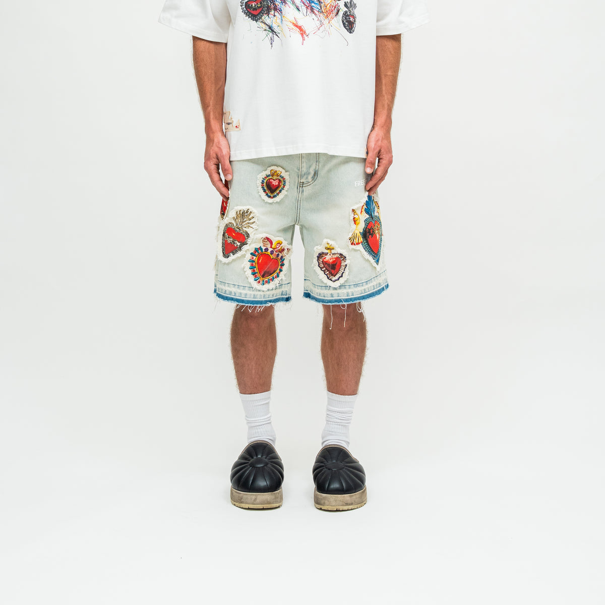 A model is shown from the waist down wearing light-wash denim shorts heavily decorated with colorful, detailed religious heart patches, paired with a white graphic t-shirt, white socks, and black espadrille platform shoes against a plain white studio background.