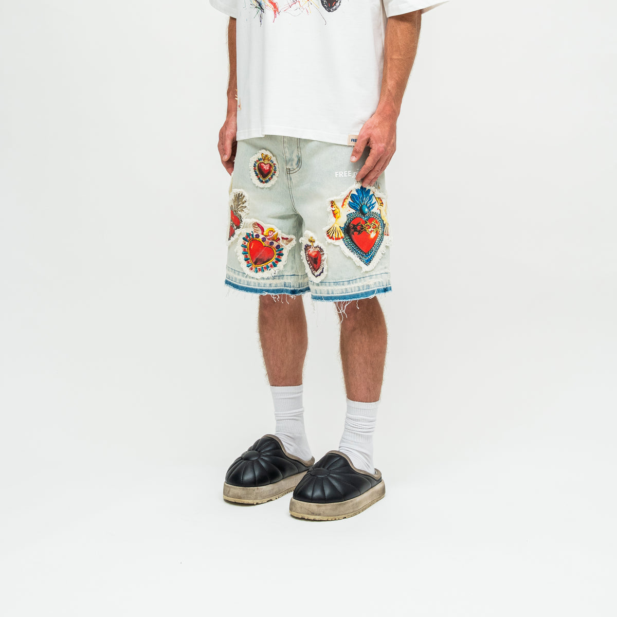 A person wearing light-wash denim shorts with colorful embroidered heart and floral motifs, a white graphic T-shirt, white socks, and black espadrille-style slip-on shoes, photographed against a clean white studio background.