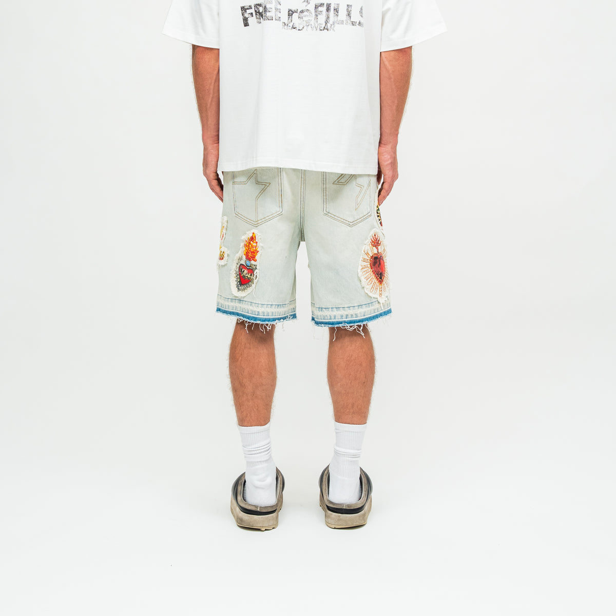 A model shown from the back wearing light-wash denim shorts with colorful embroidered patches on the back pockets and frayed hems, styled with a white graphic t-shirt, white socks, and black slide sandals against a plain white studio background.