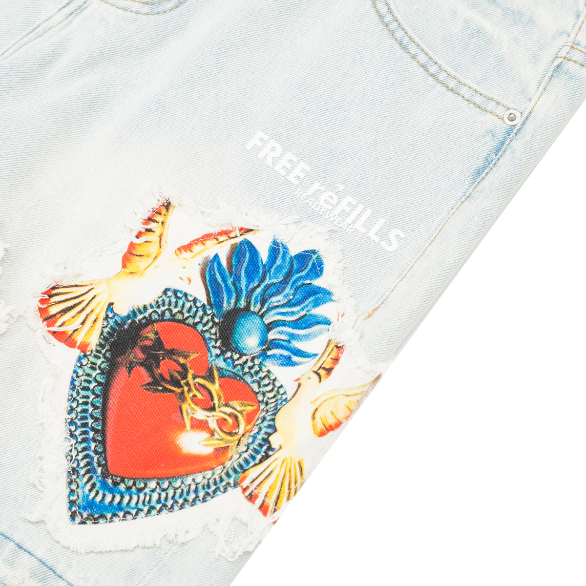 Close-up of light-wash denim jeans with a distressed patch featuring a red heart, blue flower, and gold chain details, plus white 'FREE REFILLS' text on the fabric.