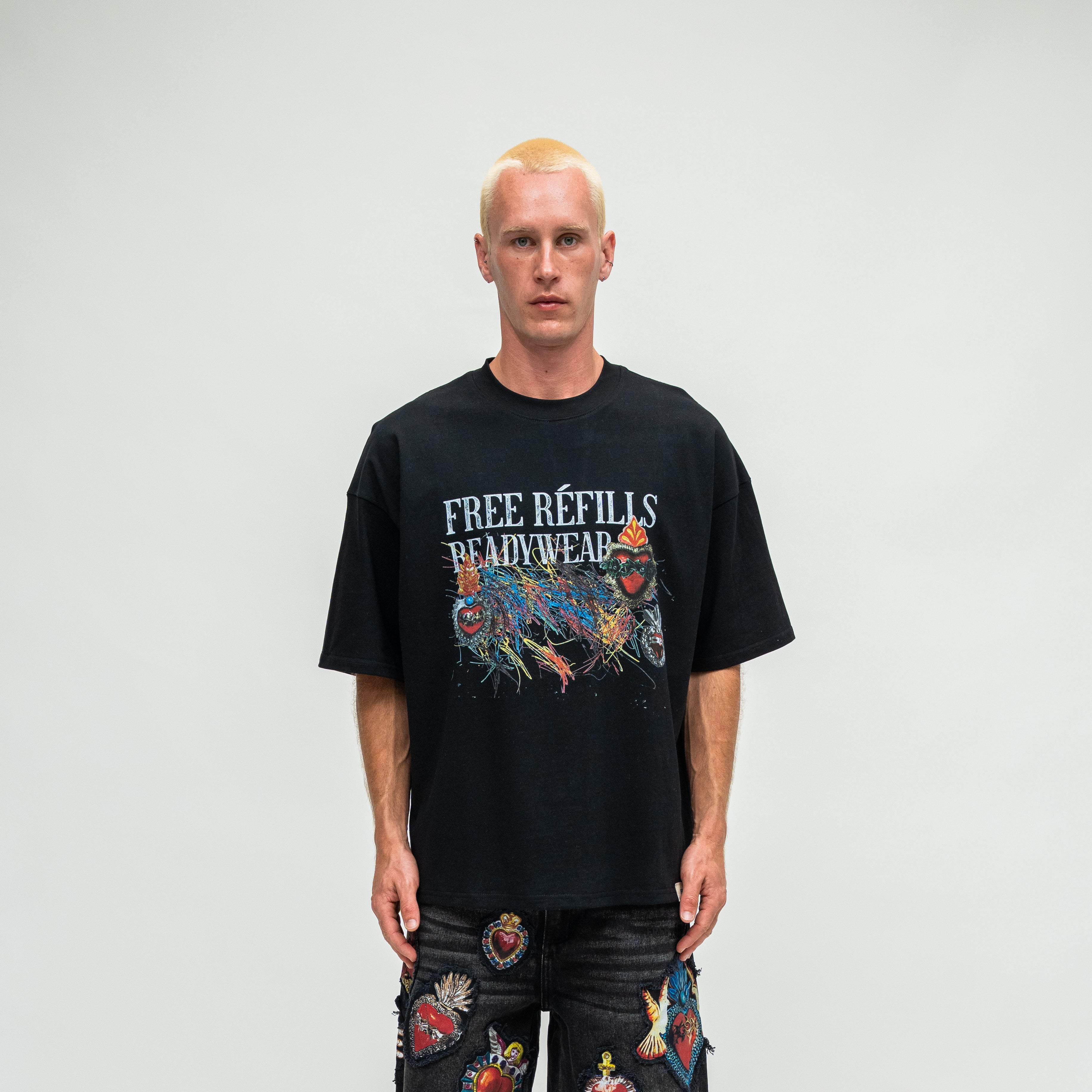 A man with short blonde hair stands against a light gray studio background wearing an oversized black graphic T-shirt with the text 'FREE REFILLS READYWEAR' and a colorful graffiti-style illustration, paired with dark jeans covered in patches.