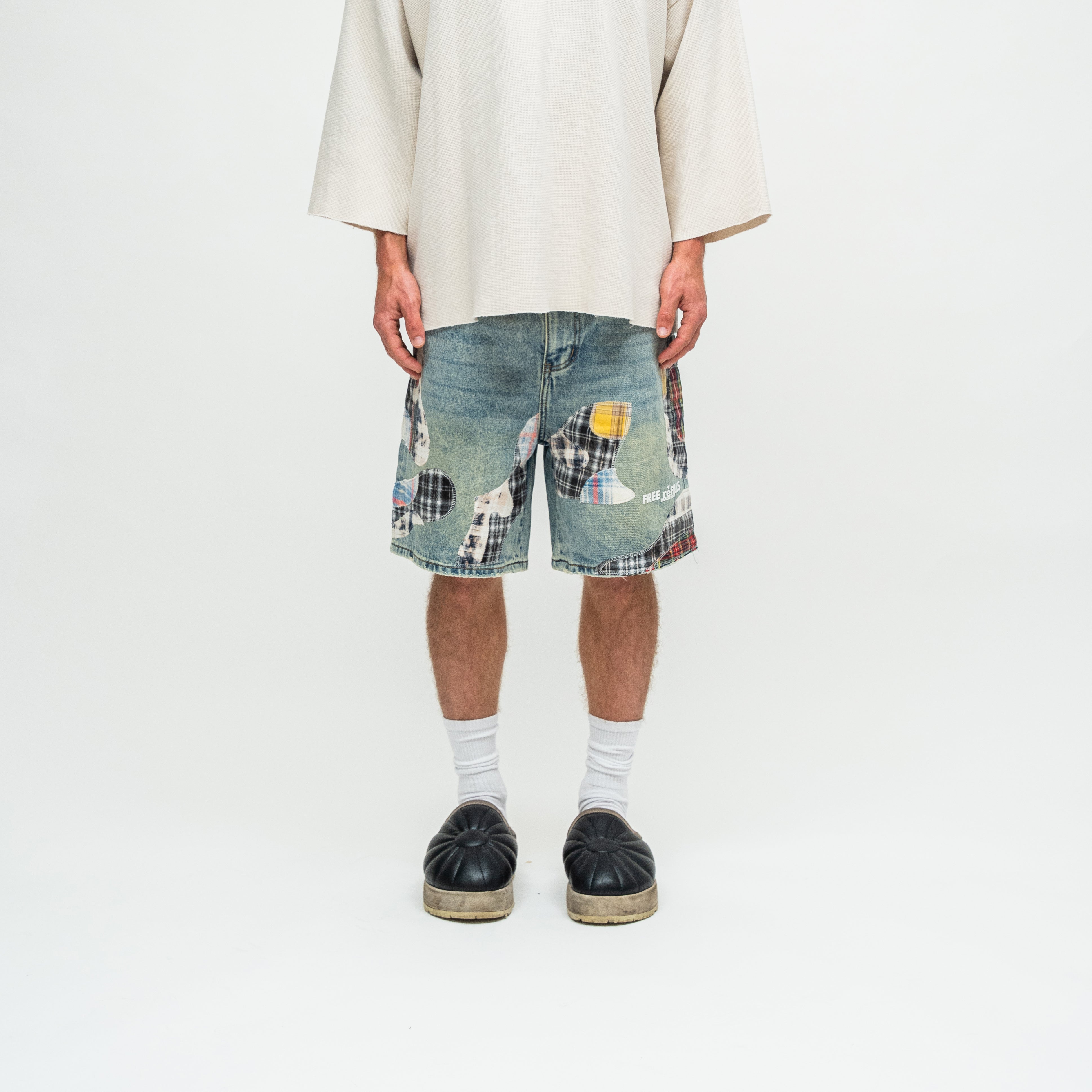 A person shown from the chest down wearing distressed denim shorts with a patchwork design, a cream-colored oversized t-shirt, white socks, and black platform sandals against a plain white studio background.