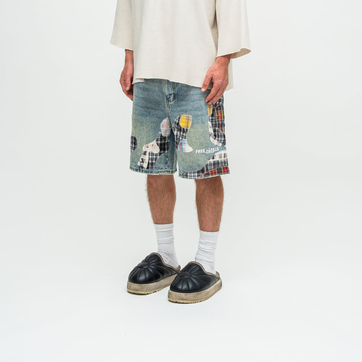 A person wearing light-wash denim shorts with a patchwork design featuring tartan plaid and a white dog illustration, styled with white crew socks and black espadrille slip-on shoes against a plain white studio background.