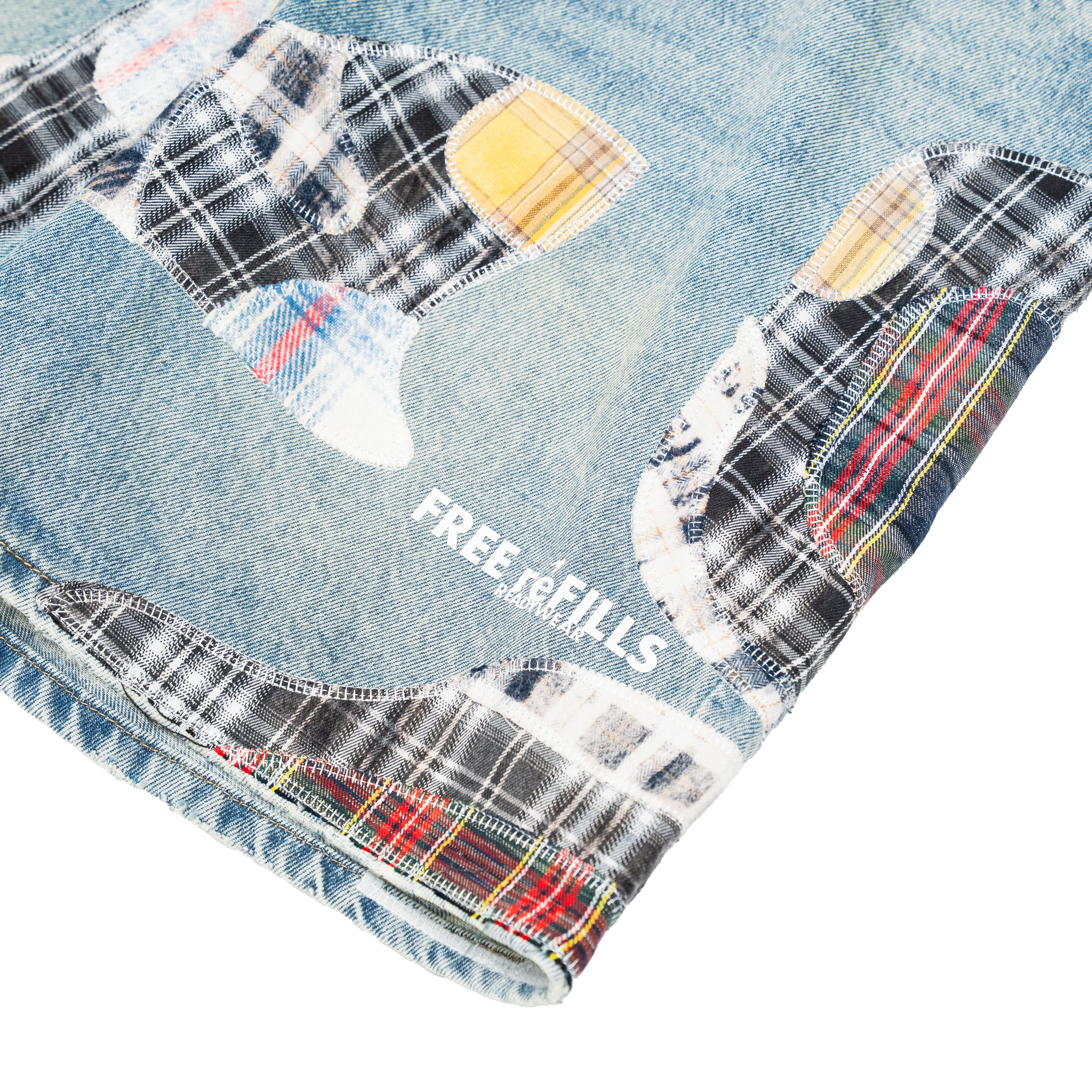Close-up of a light blue denim garment with large patchwork plaid patches in black, white, red, yellow, and blue, plus the visible text 'FREE REFILLS' on a white background.