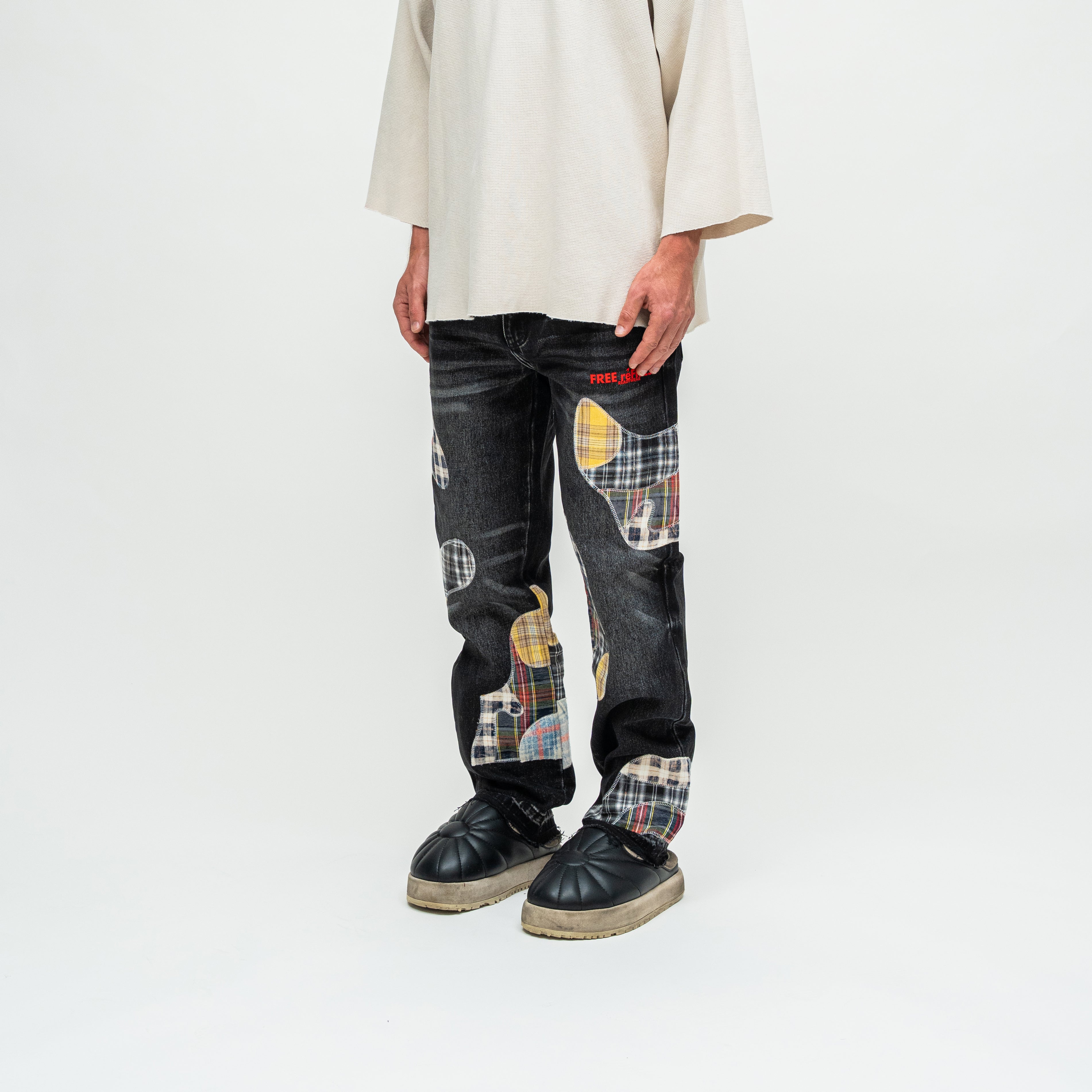 A person wearing dark patchwork denim jeans with colorful tartan and circular patch details, paired with a cream-colored shirt and black shoes, photographed against a plain white studio background.