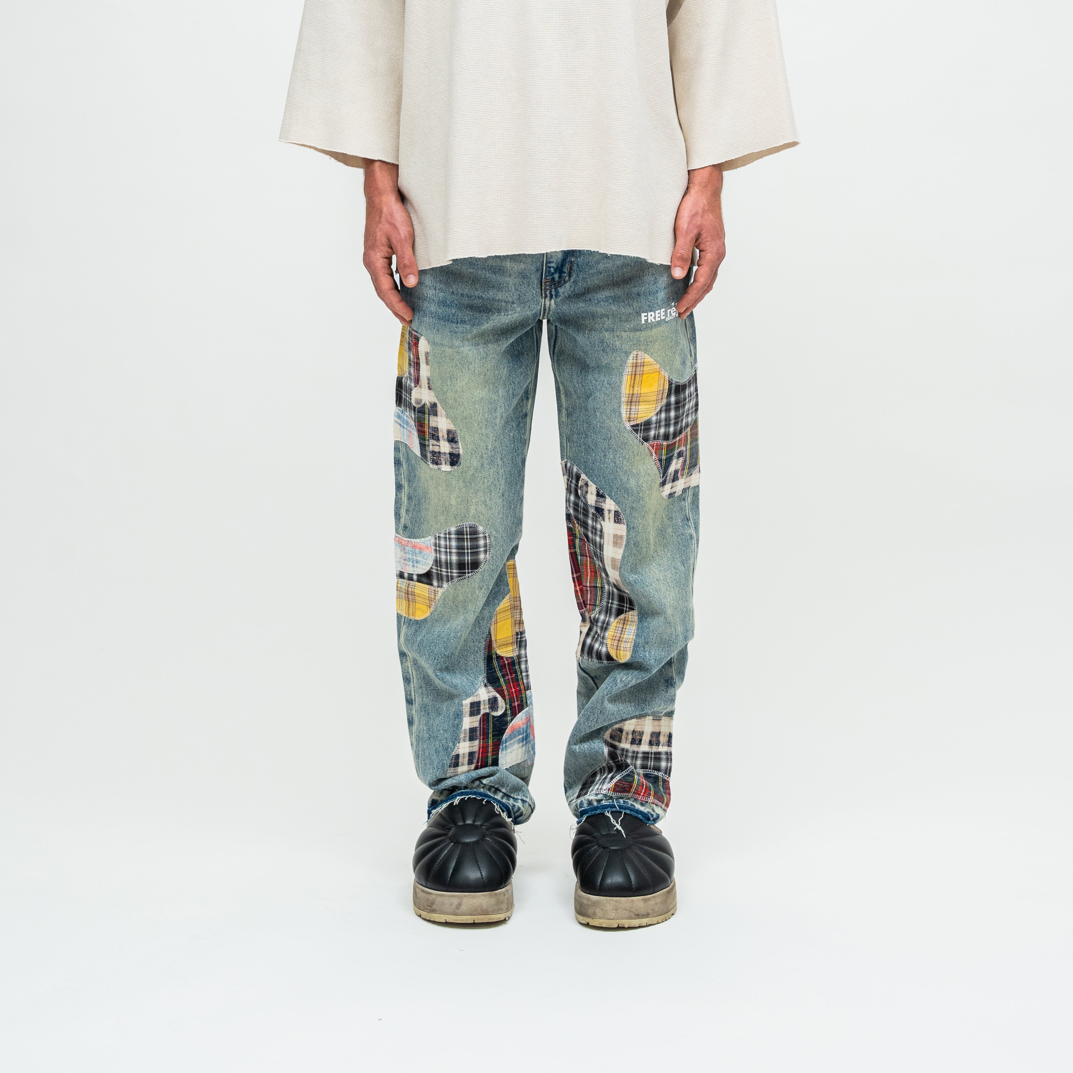 A person wearing loose-fit blue denim jeans with large multicolor patchwork plaid inserts, paired with a cream shirt and black shoes, photographed against a plain white studio background.