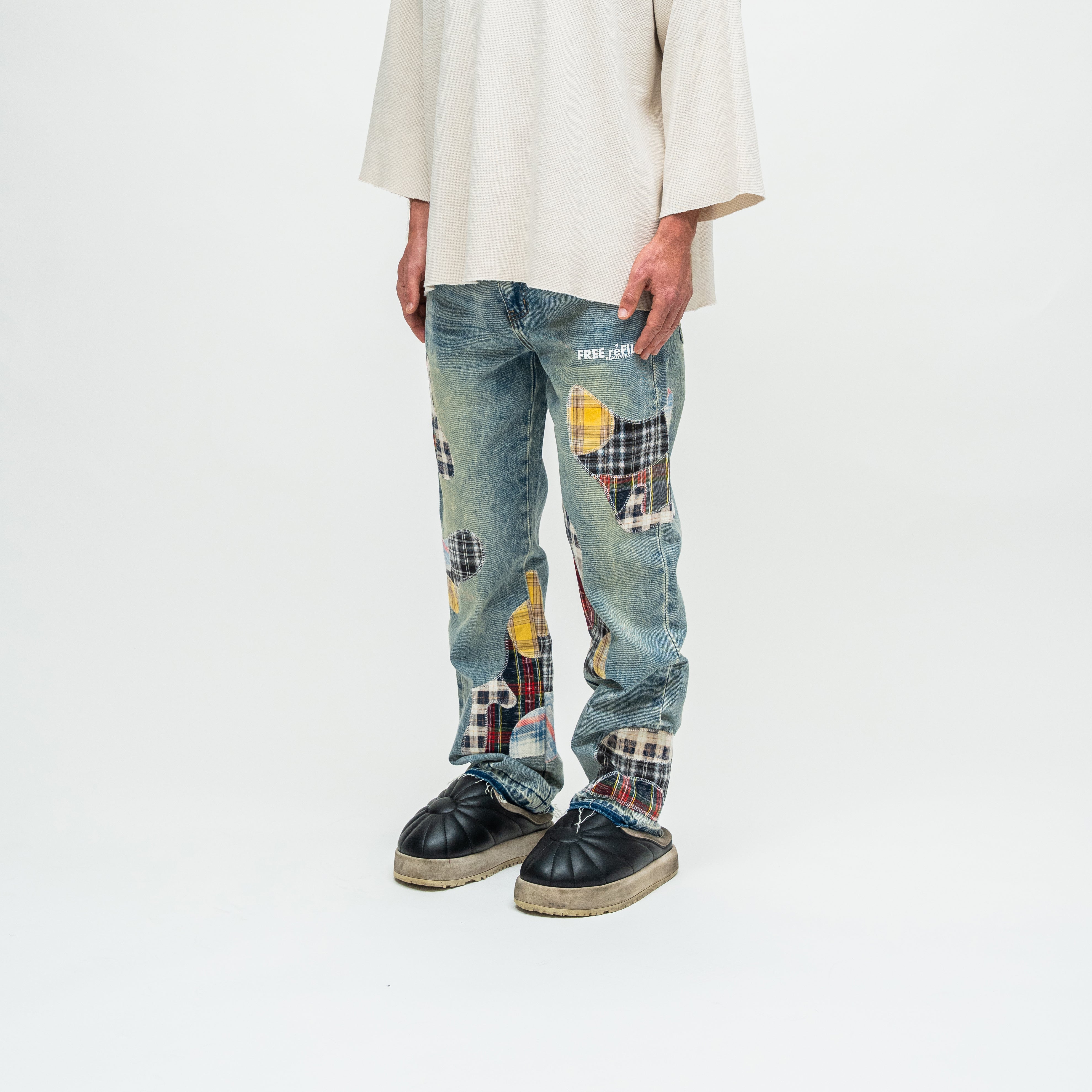 A person wearing light blue patchwork denim jeans with yellow, red, and black plaid details, a cream-colored oversized top, and black leather shoes, photographed against a plain white studio background.