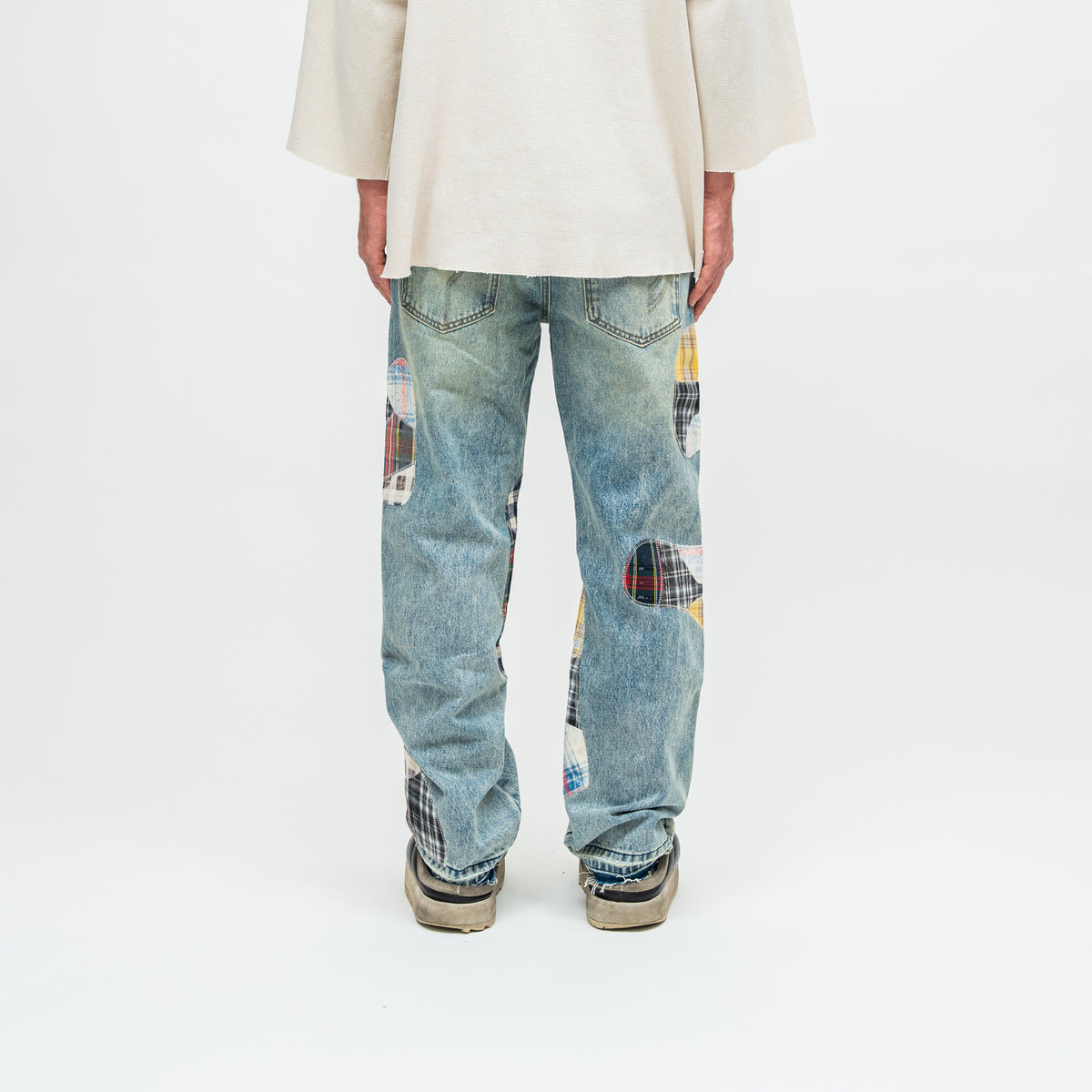 A person shown from the back wearing light blue patchwork denim jeans with colorful plaid inserts, a cream-colored top, and beige sneakers against a plain white studio background.