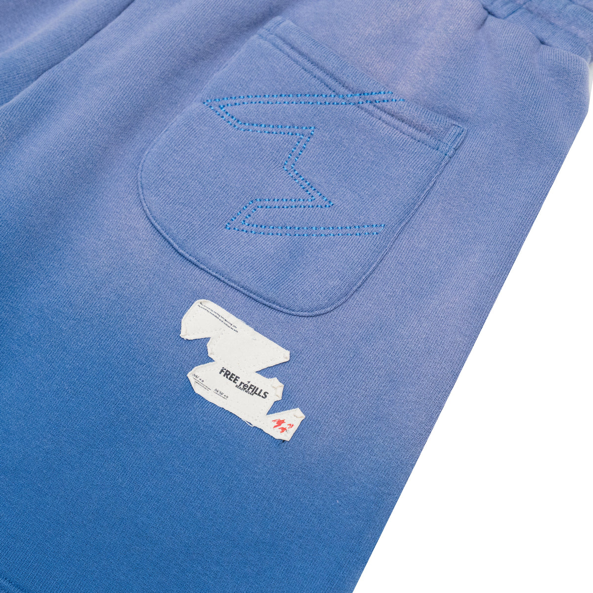 Close-up of blue denim shorts showing a back pocket with decorative blue zigzag stitching and a torn white paper patch with the text 'FREE REFILLS' and a small red logo.