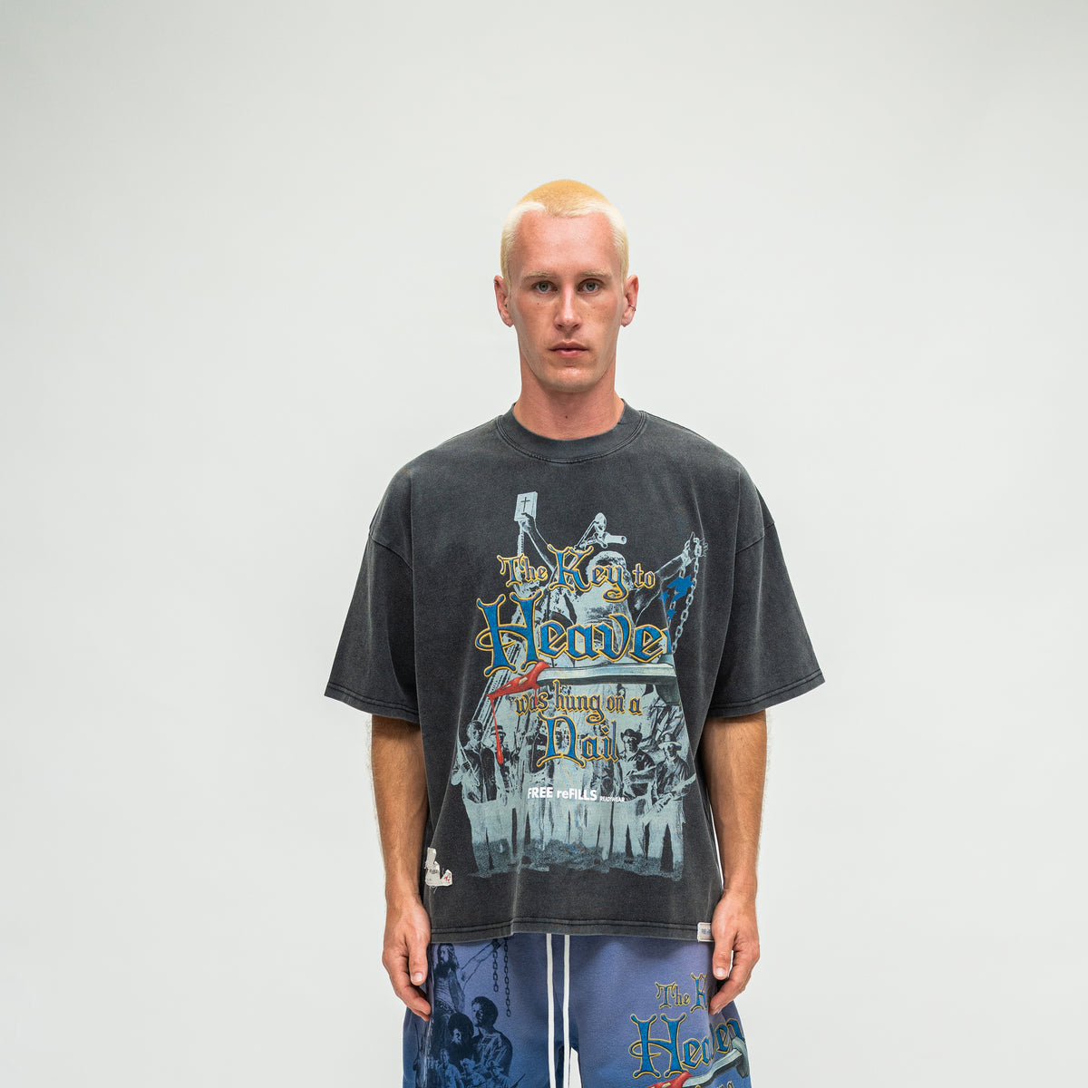 A man wearing a faded black graphic t-shirt with a vintage-style illustration and text, paired with blue shorts, standing against a plain light gray background.