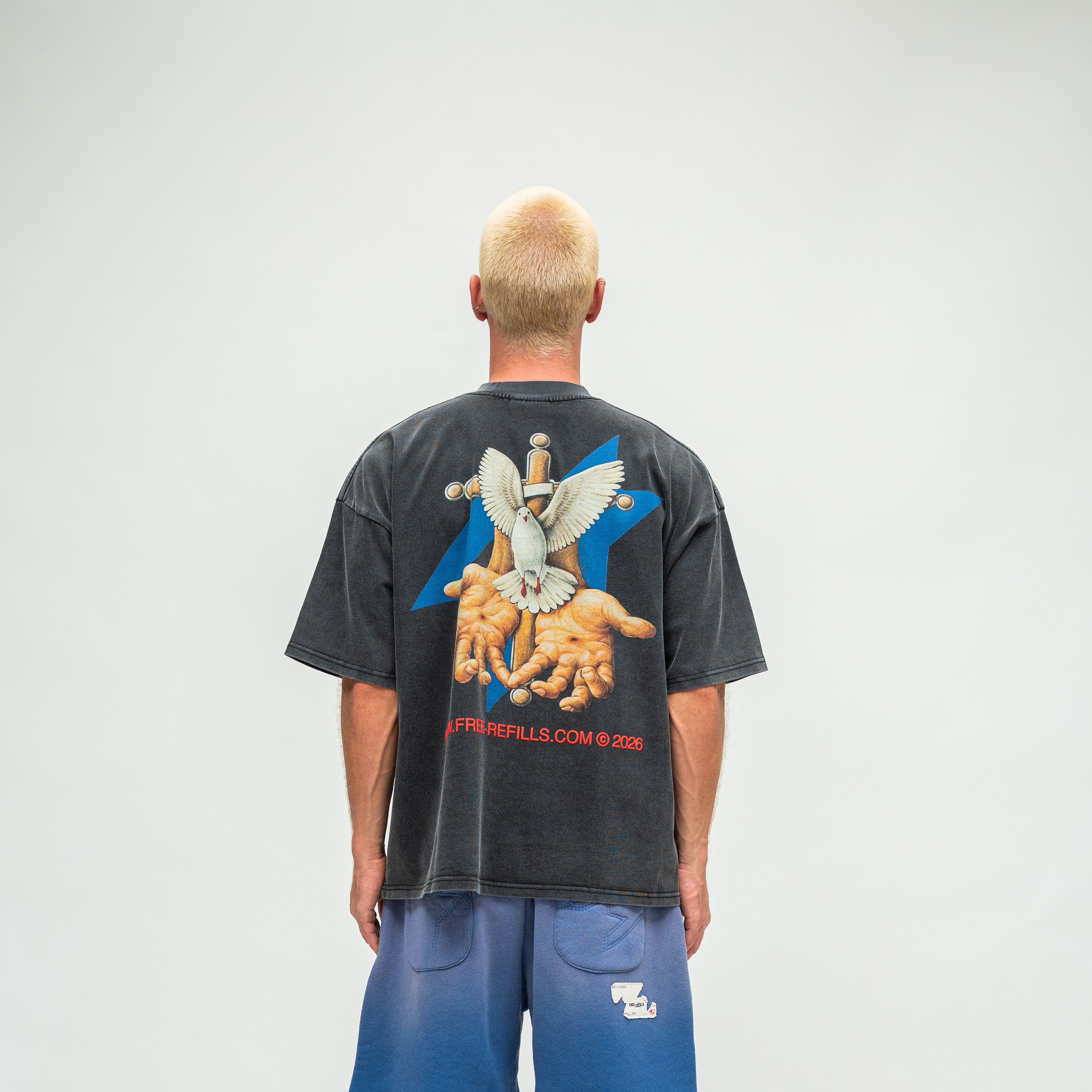 A man viewed from behind wearing a faded black graphic t-shirt and blue sweatpants against a light gray studio background.