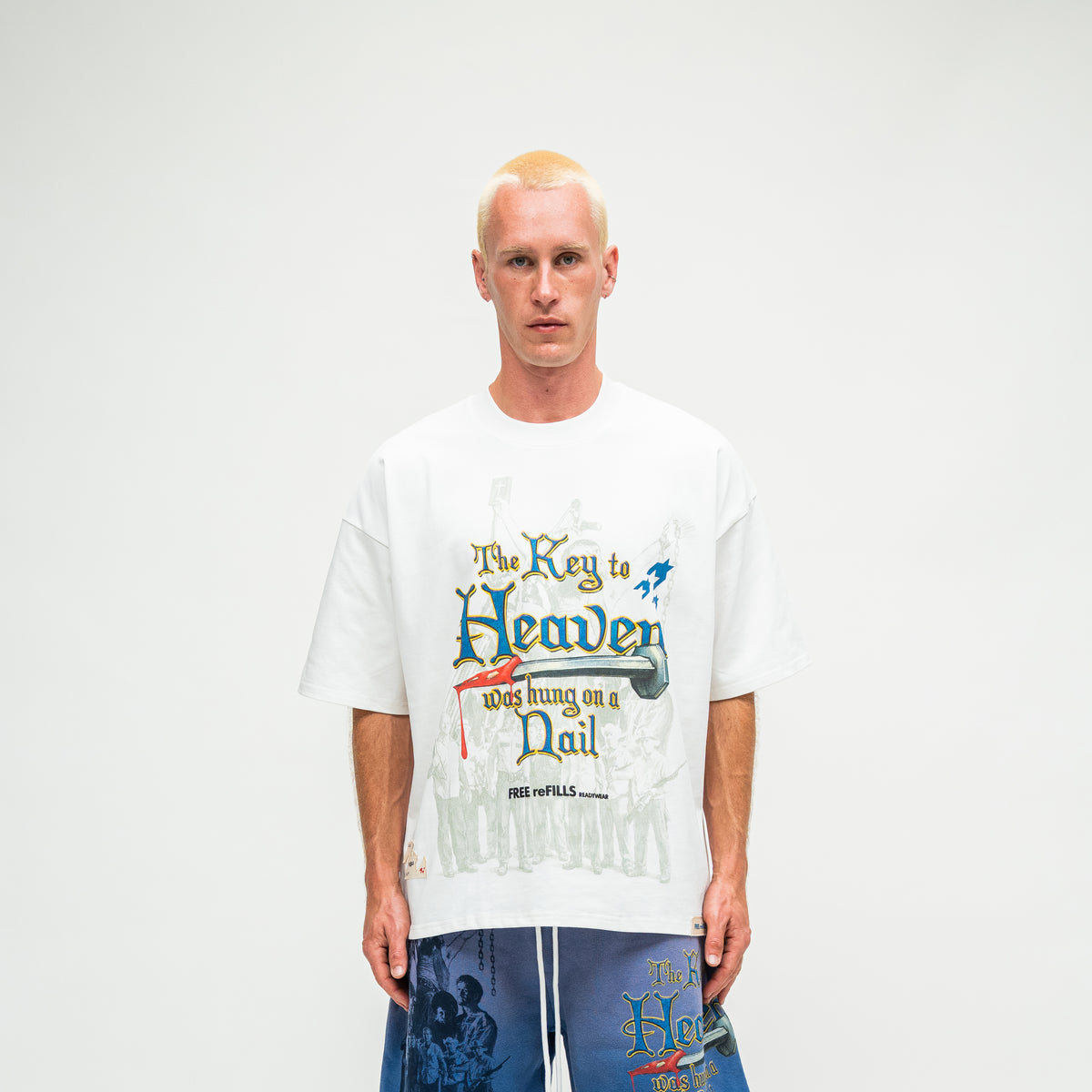 A man wearing a white graphic t-shirt with 'The Key to Heaven was hanging on a Nail' text and 'FREE REFILLS' branding, paired with blue patterned shorts, against a plain white background.