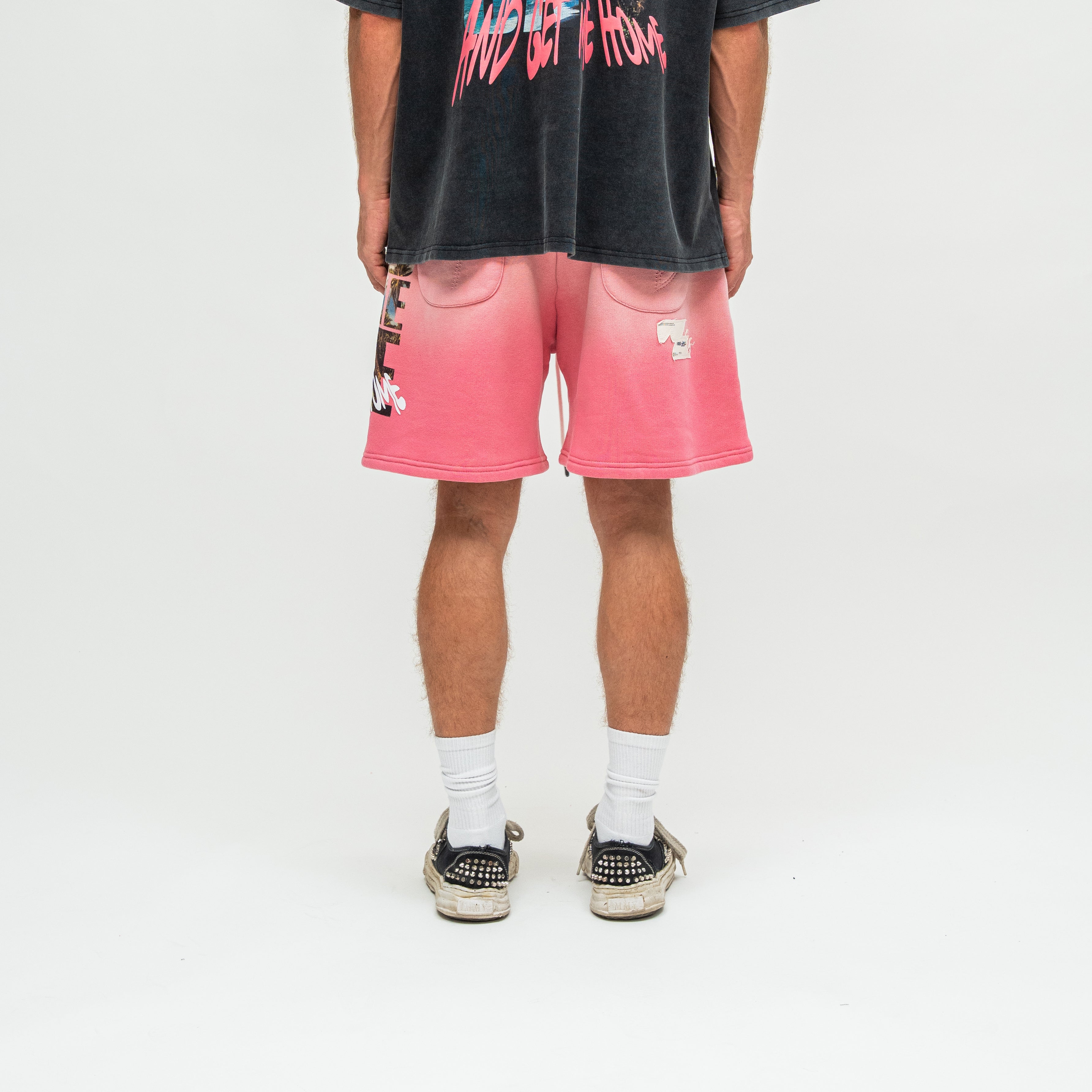 A person shown from the back wearing pink gradient sweat shorts with graphic details, a faded black graphic t-shirt, white socks, and black studded sneakers on a white studio background.