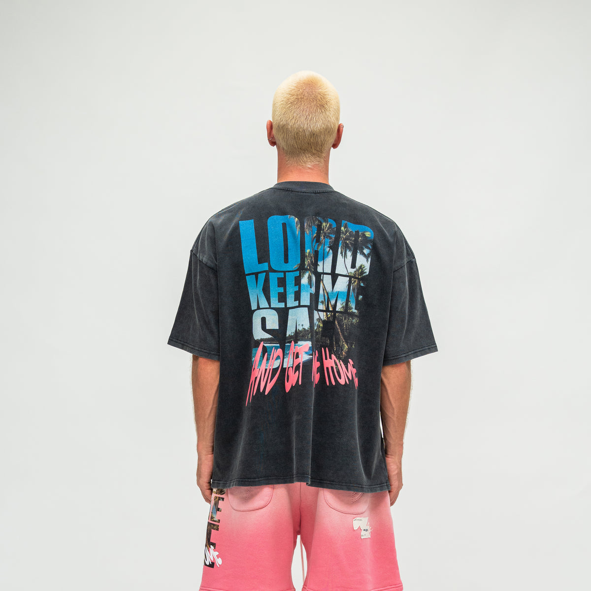 A man viewed from behind wearing a faded black graphic t-shirt with blue and pink text on the back, paired with pink shorts, against a plain light gray background.