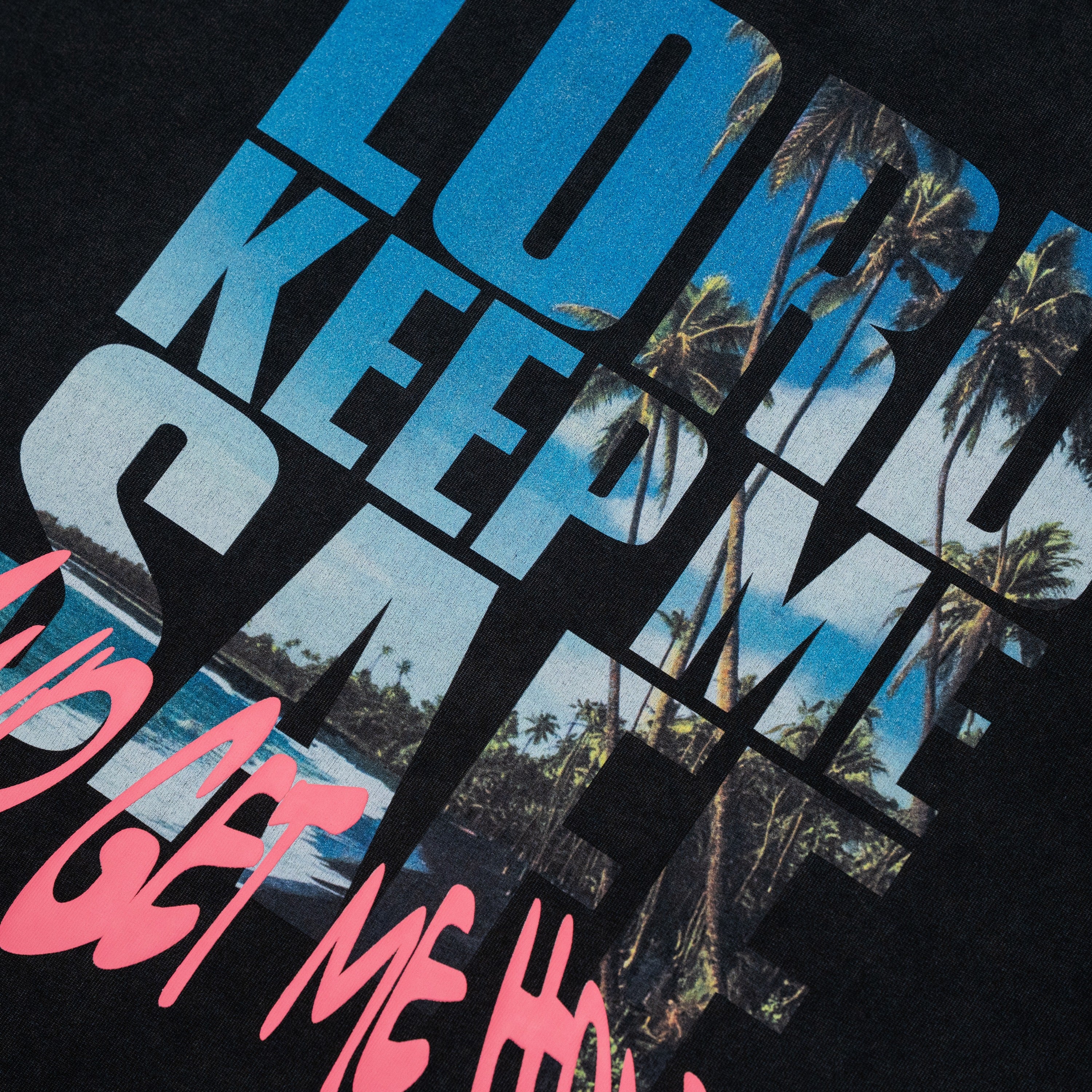 Close-up of a black graphic T-shirt print featuring large blue and white text reading 'LOVE KEEP ME SAFE' over a tropical palm tree scene, with pink text reading 'AND GET ME HOME' at the bottom.
