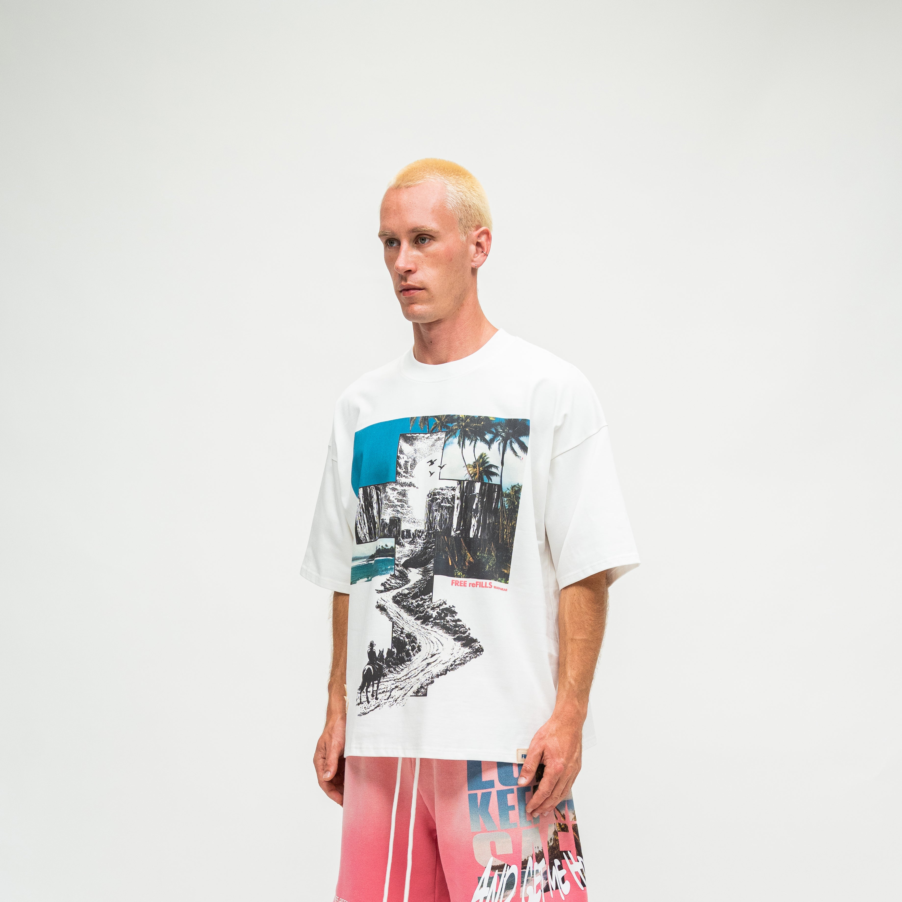 A man with blonde hair wearing a white oversized graphic t-shirt and pink shorts, standing against a plain light gray studio background.