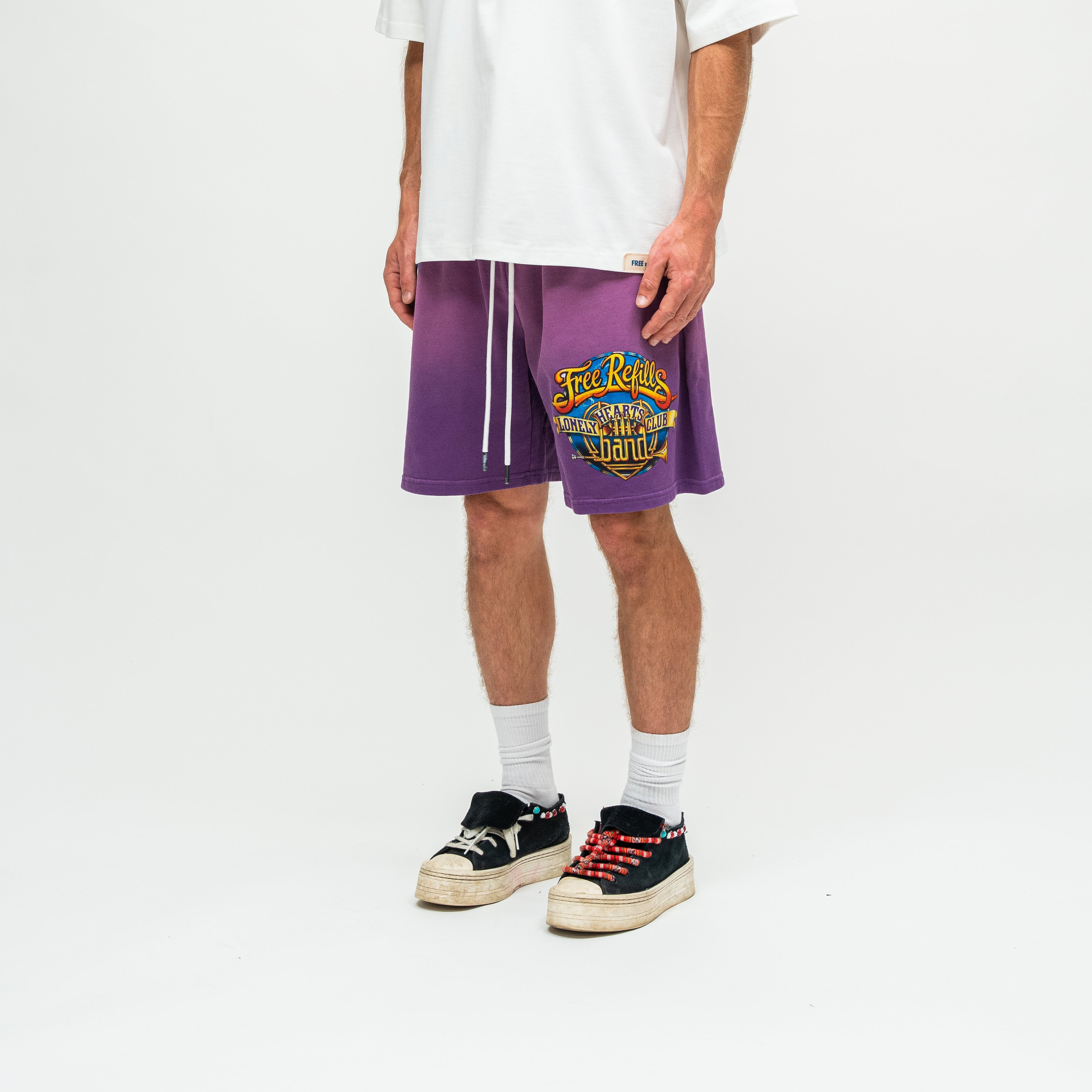 A person wearing purple drawstring shorts with a colorful 'FREE REFILLS' graphic on the left leg, styled with a white t-shirt, white socks, and black low-top sneakers on a white studio background.