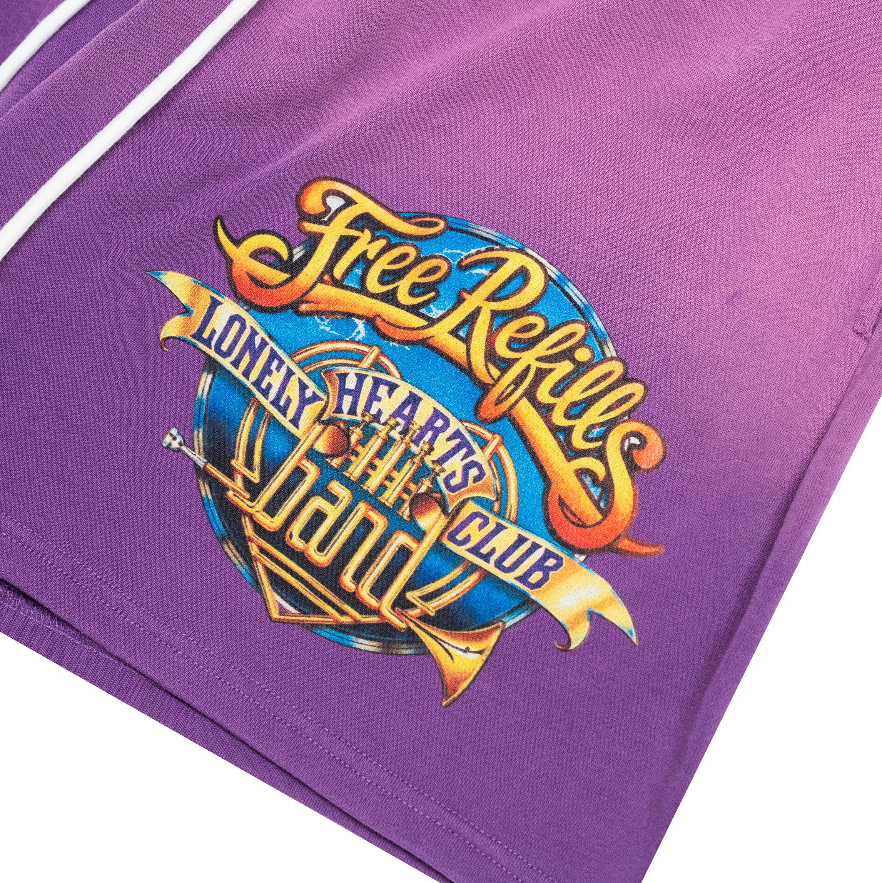 Close-up of a purple garment, likely shorts or a sweatshirt, featuring a large colorful graphic print on the front. The graphic reads 'Free Refills' and 'LONELY HEARTS CLUB' in stylized yellow, orange, and blue lettering.