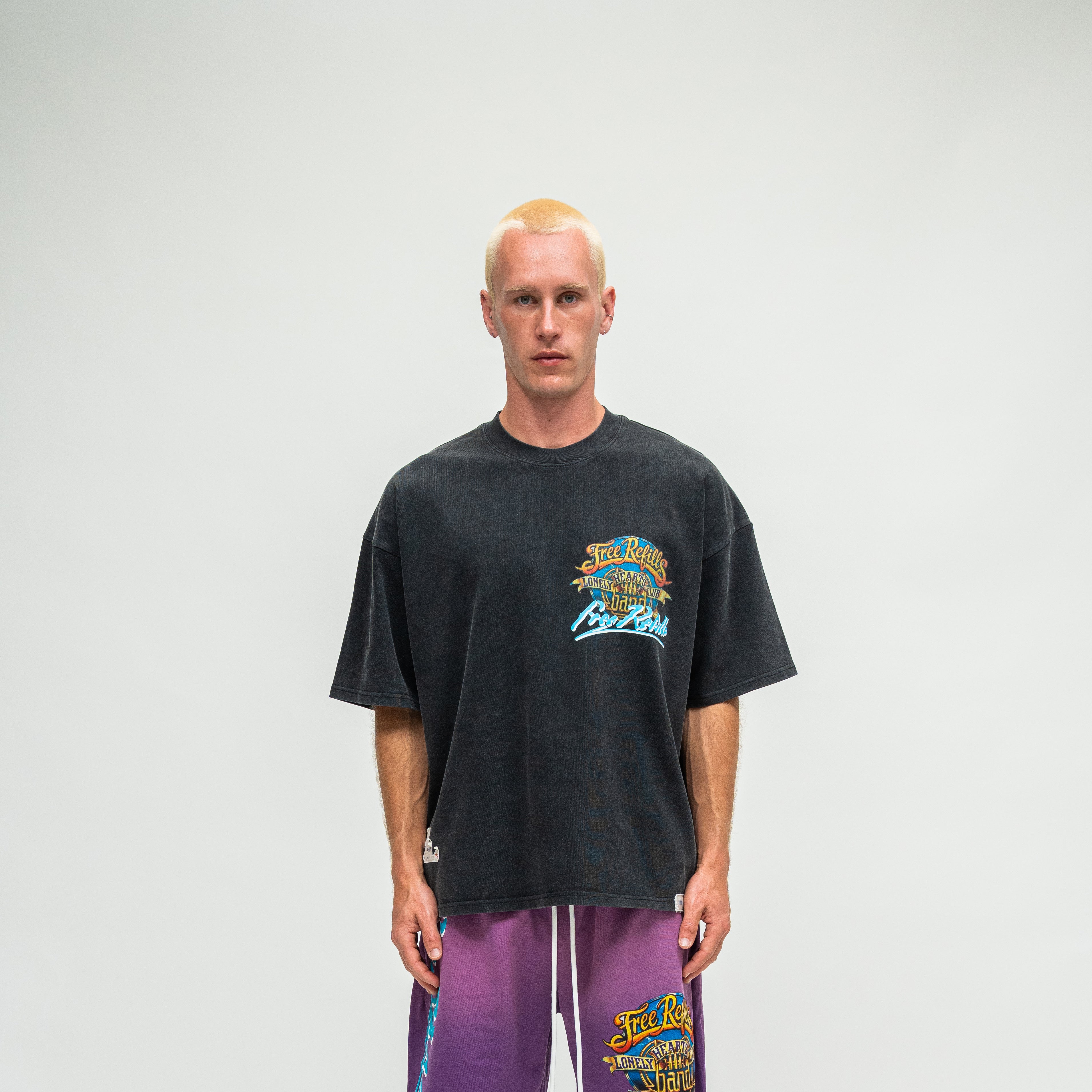 A man wearing a faded black graphic t-shirt and purple sweatpants, standing against a light gray studio background.