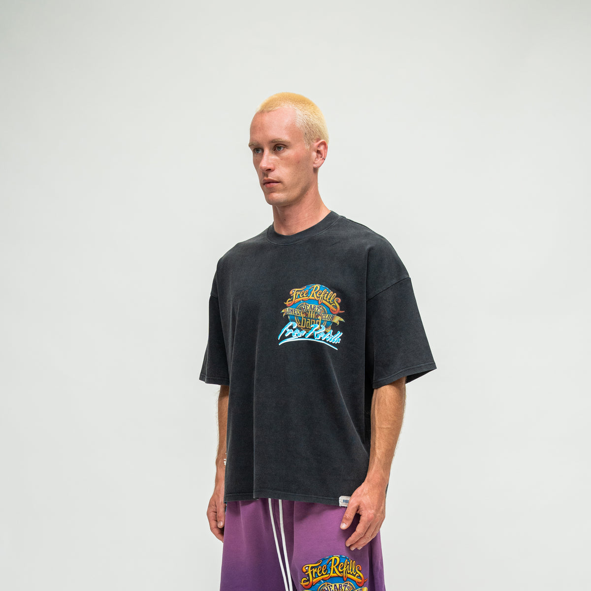 A man wearing an oversized black washed graphic t-shirt and purple shorts, standing against a light gray studio background.