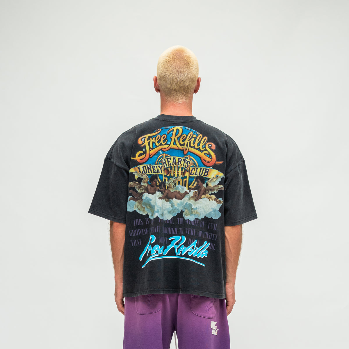 A man viewed from behind wearing a faded black graphic t-shirt with a colorful 'Free Refills' print and purple pants, set against a plain light gray background.