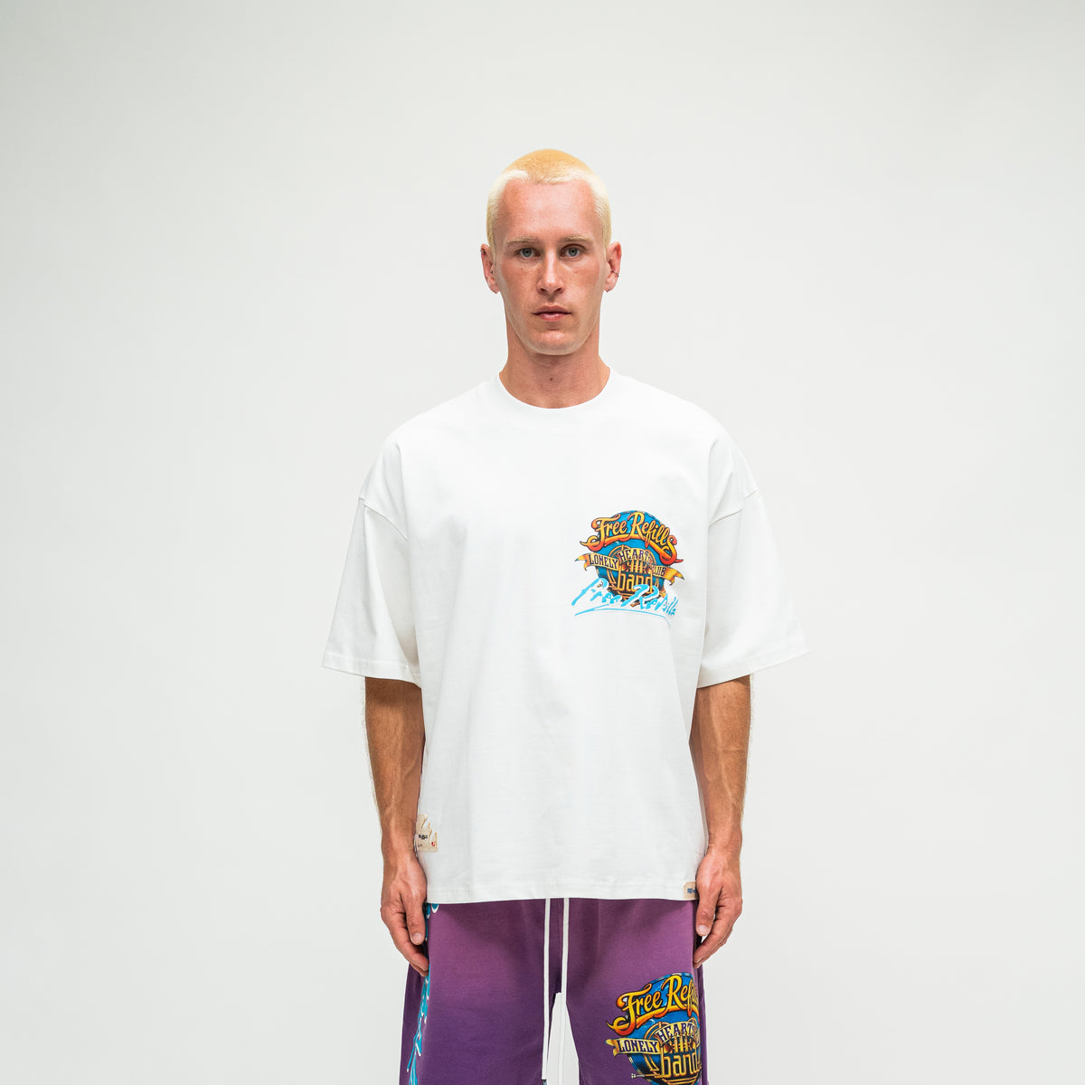 A man with a buzz cut stands against a light gray studio background wearing a white short-sleeve t-shirt with a colorful graphic on the left chest and purple drawstring sweatpants with a matching graphic.