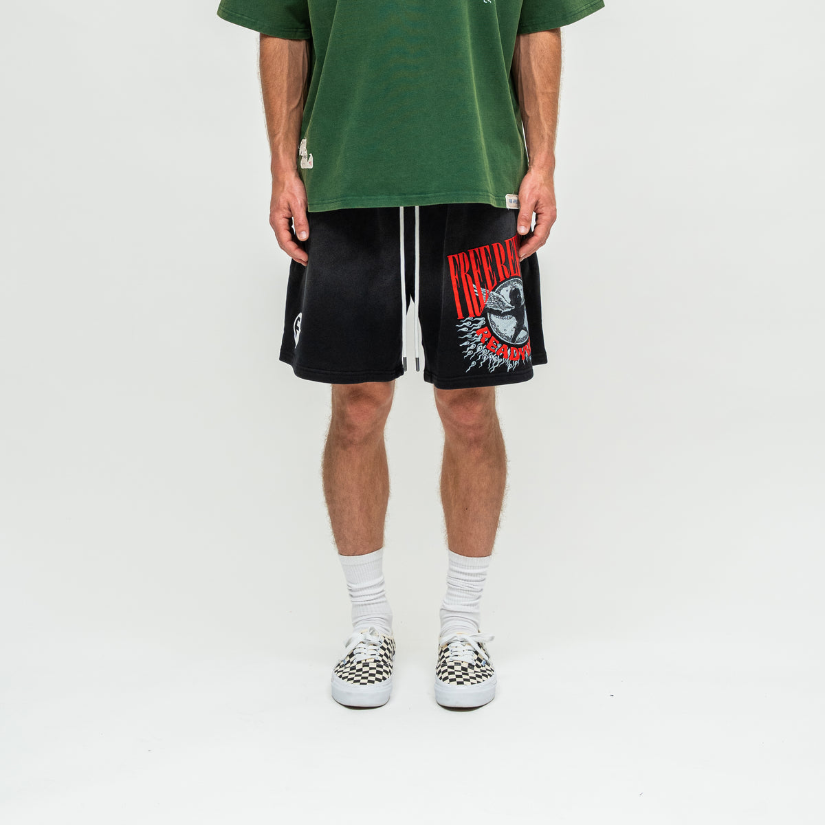 A person wearing black graphic athletic shorts with a red and white 'FREE REFILLS' design, a green t-shirt, white socks, and checkerboard sneakers, standing against a light gray studio background.