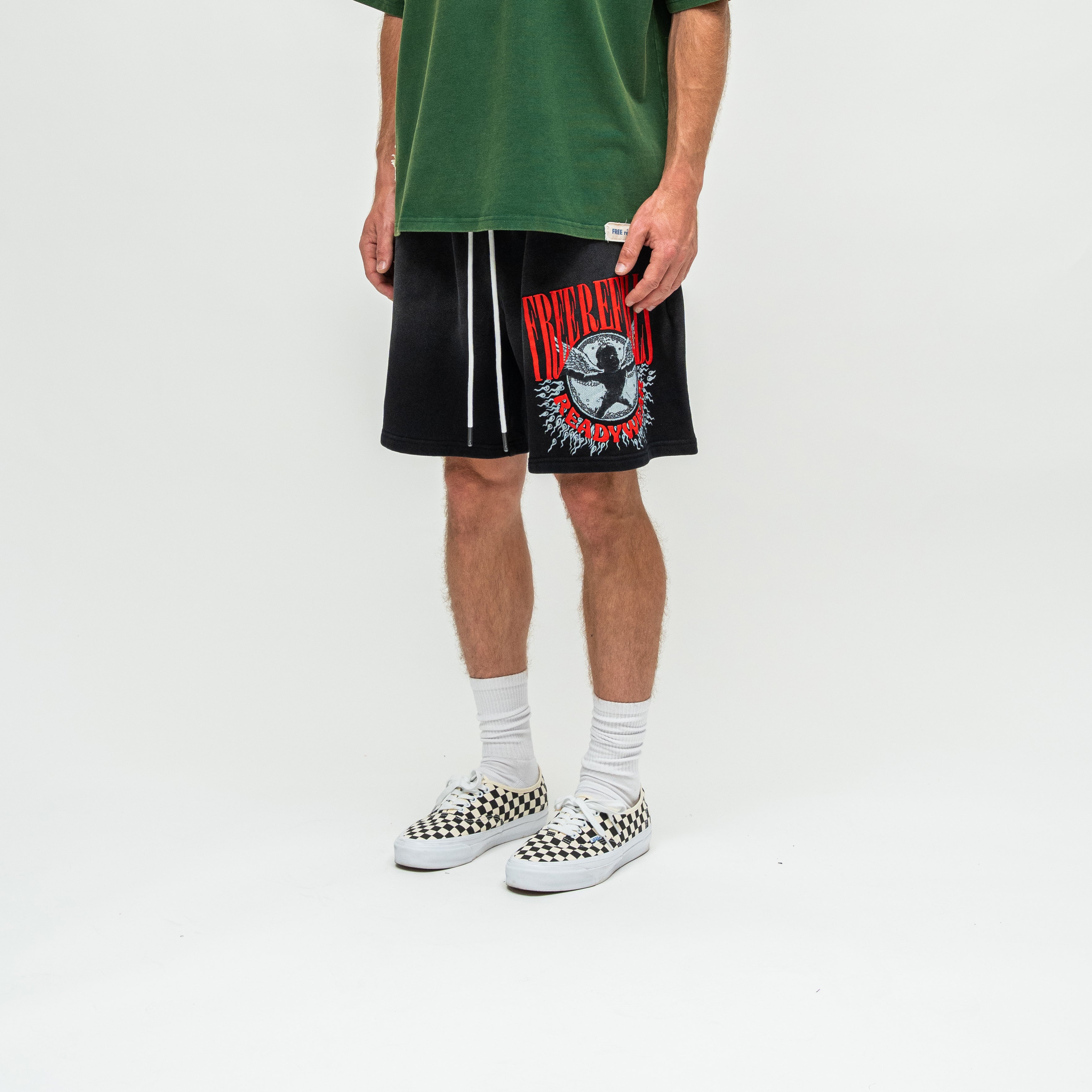 A person wearing black athletic shorts with a red and white graphic print, a green t-shirt, white socks, and black-and-white checkered Vans sneakers, standing against a plain white studio background.