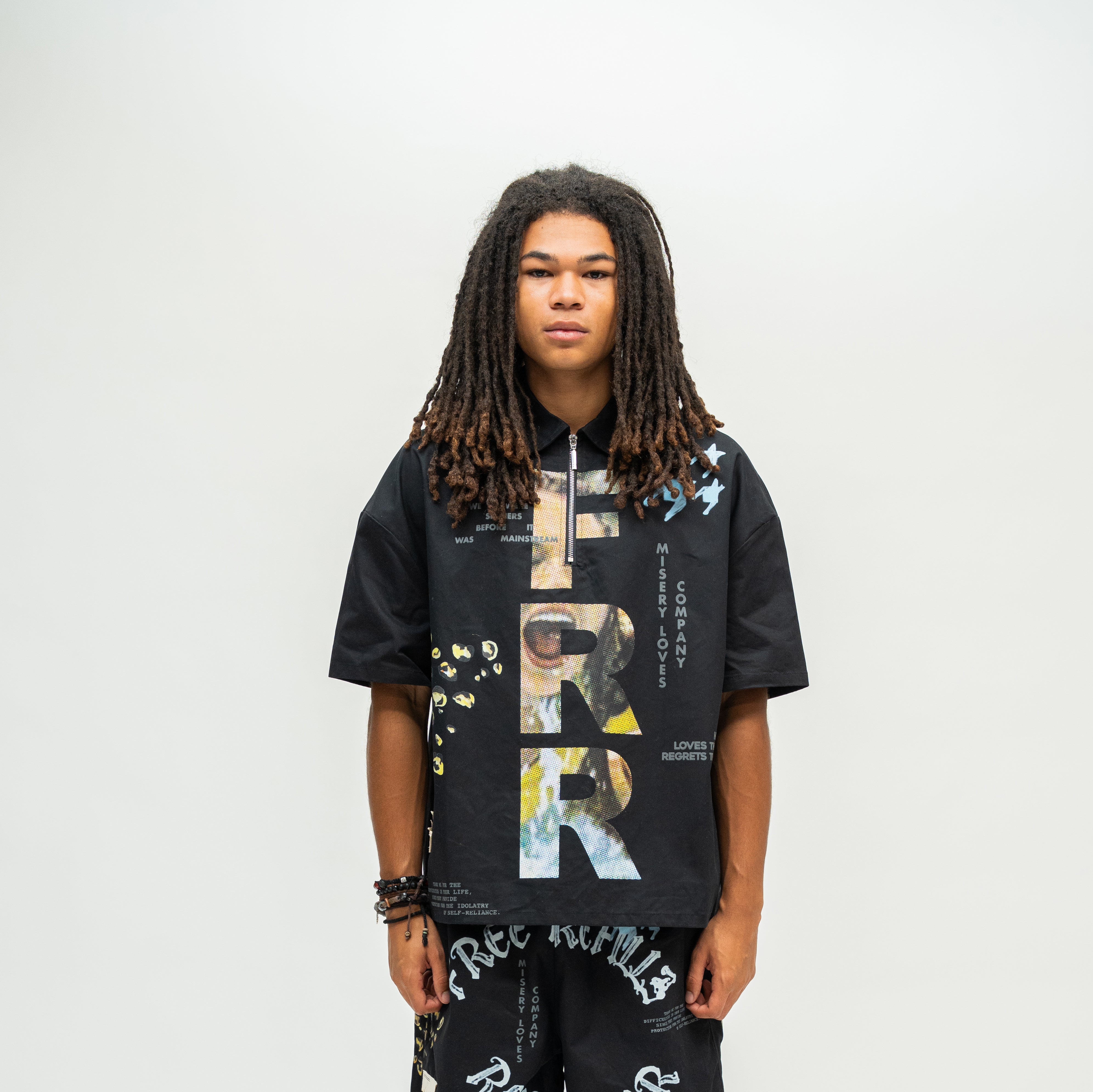 A man with long dreadlocks stands against a light gray studio background wearing a black oversized zip-up t-shirt with a large graphic print featuring the word 'FATHER' in a collage style, and matching black pants with white text.