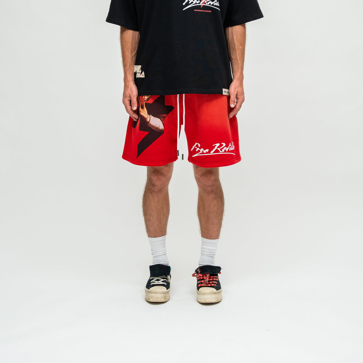 A model is shown from the waist down wearing red athletic shorts with a white drawstring and 'FREE REFILLS' branding, paired with a black t-shirt, white socks, and black sneakers with red laces, against a plain white studio background.