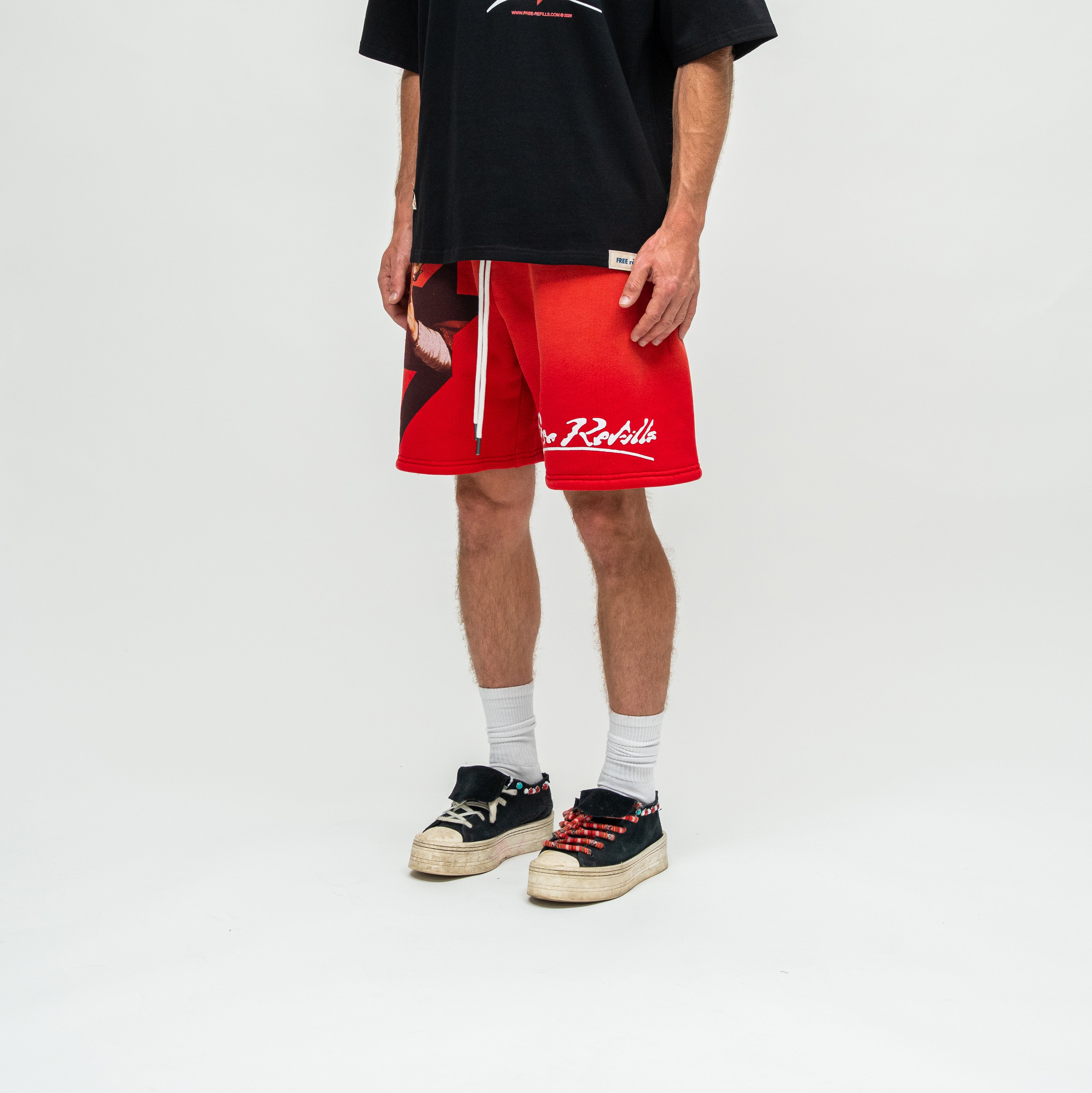 A person wearing bright red athletic shorts with white script branding and a white drawstring, styled with a black graphic T-shirt, white socks, and black low-top sneakers on a light gray studio background.