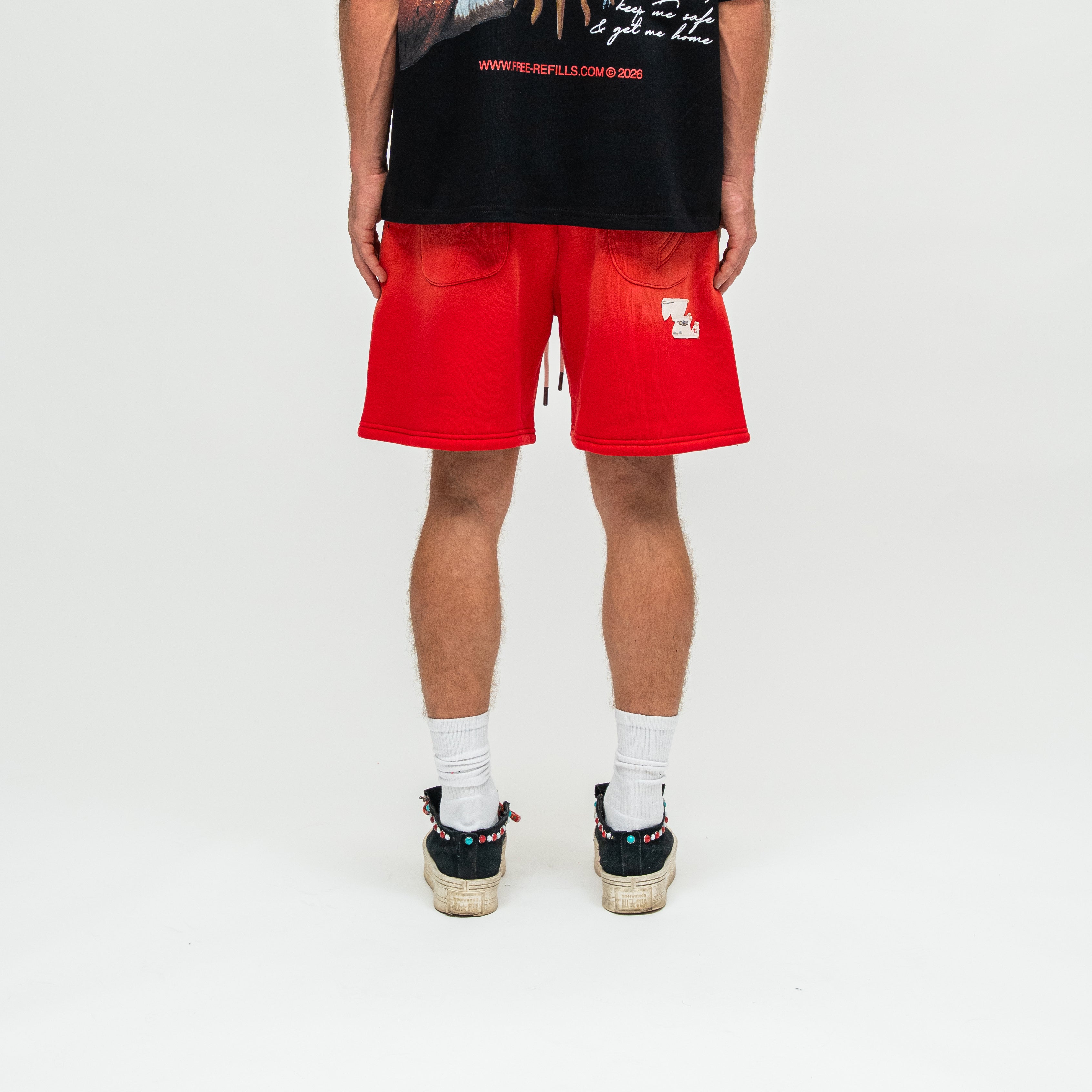 A person shown from the back wearing bright red shorts, a black graphic T-shirt, white socks, and black sneakers on a clean white studio background.