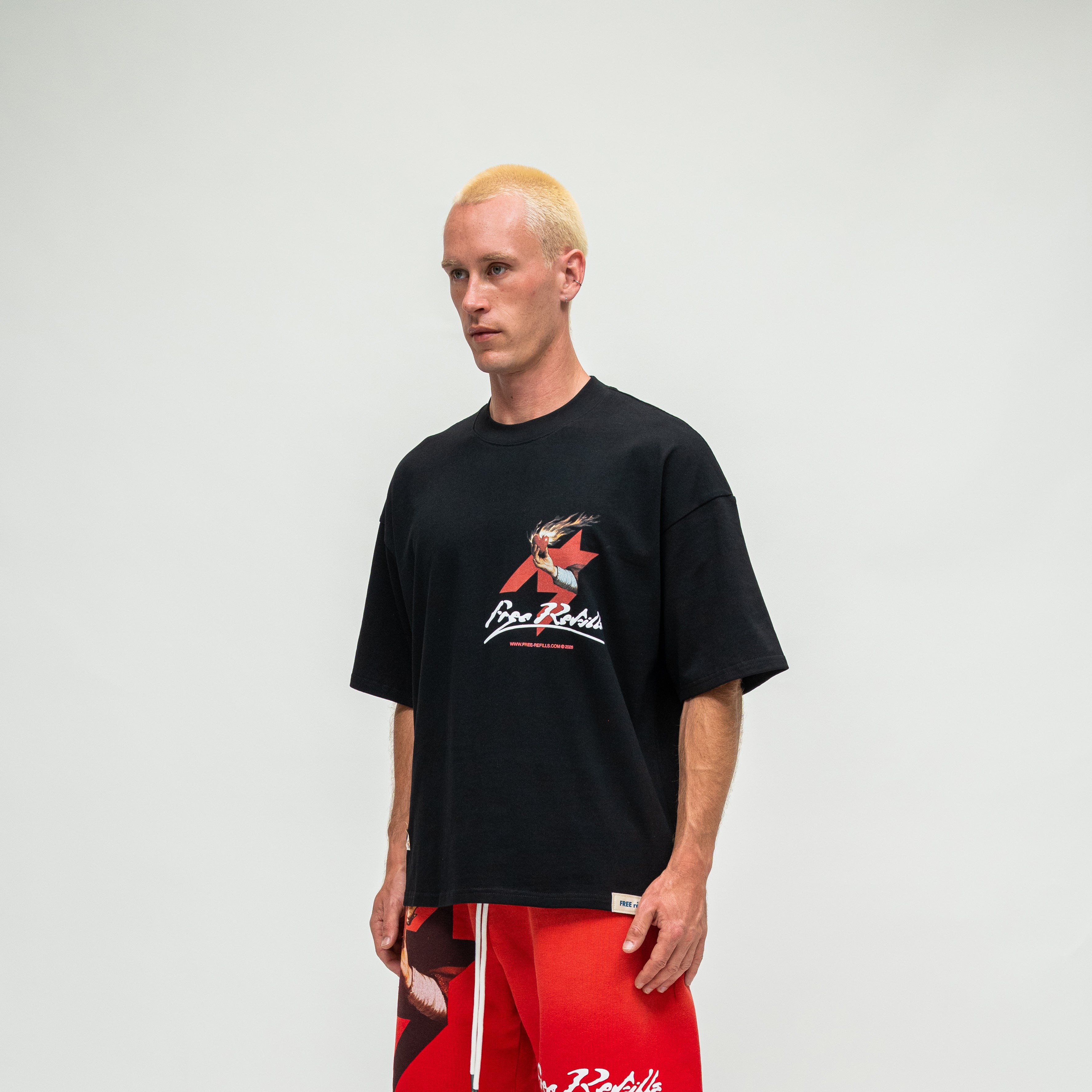 A man with blonde hair wearing a black t-shirt with a red and white graphic logo and red shorts, standing against a light gray background.