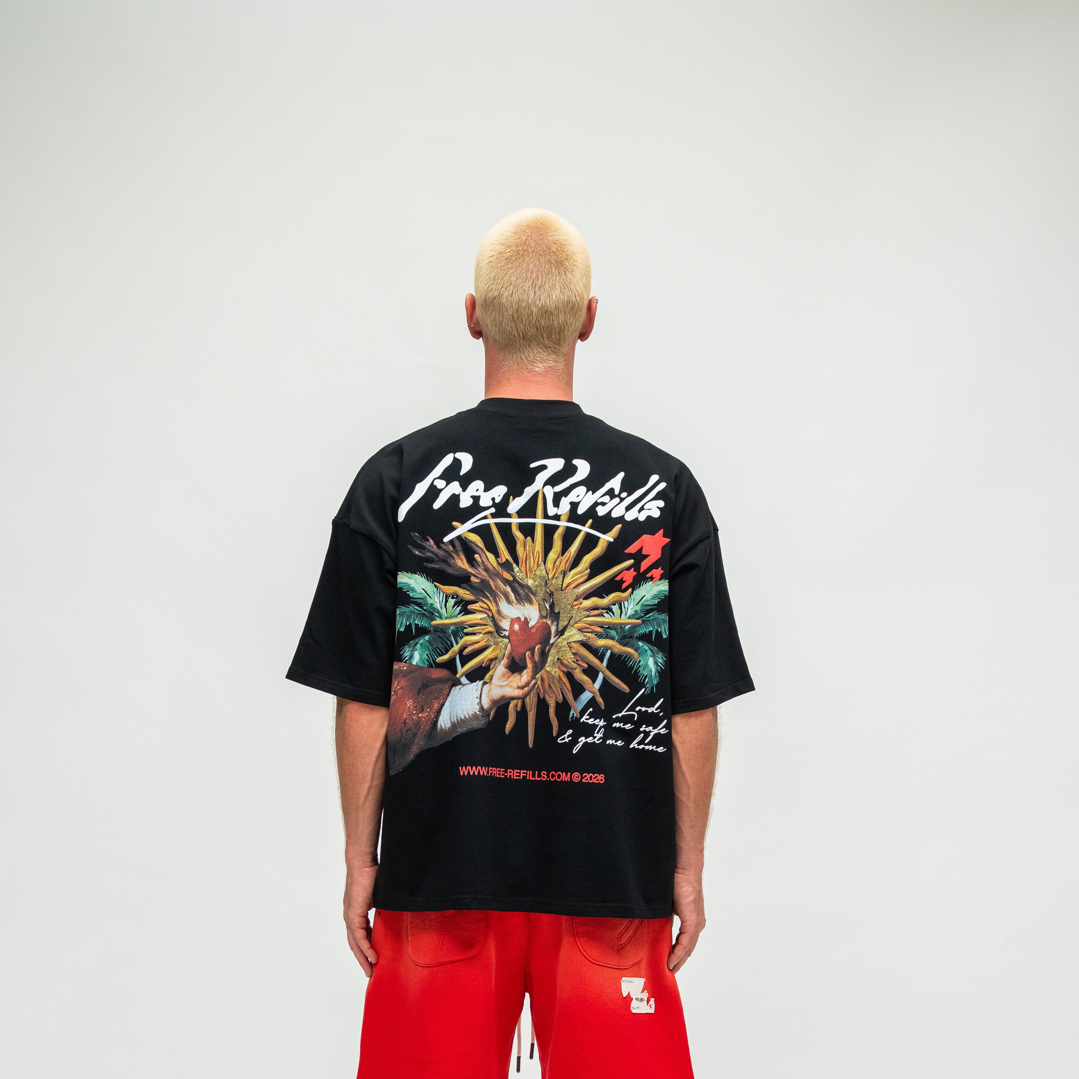 A man viewed from behind wearing a black graphic t-shirt with a large multicolor dragon design and white 'Free Refills' branding, paired with bright red shorts, against a light gray studio background.
