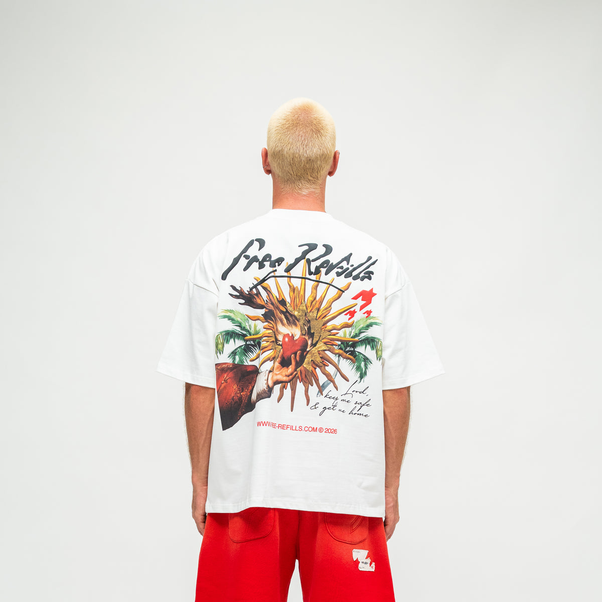 A man viewed from behind wearing a white t-shirt with a large colorful graphic print on the back and red shorts, standing against a light gray studio background.