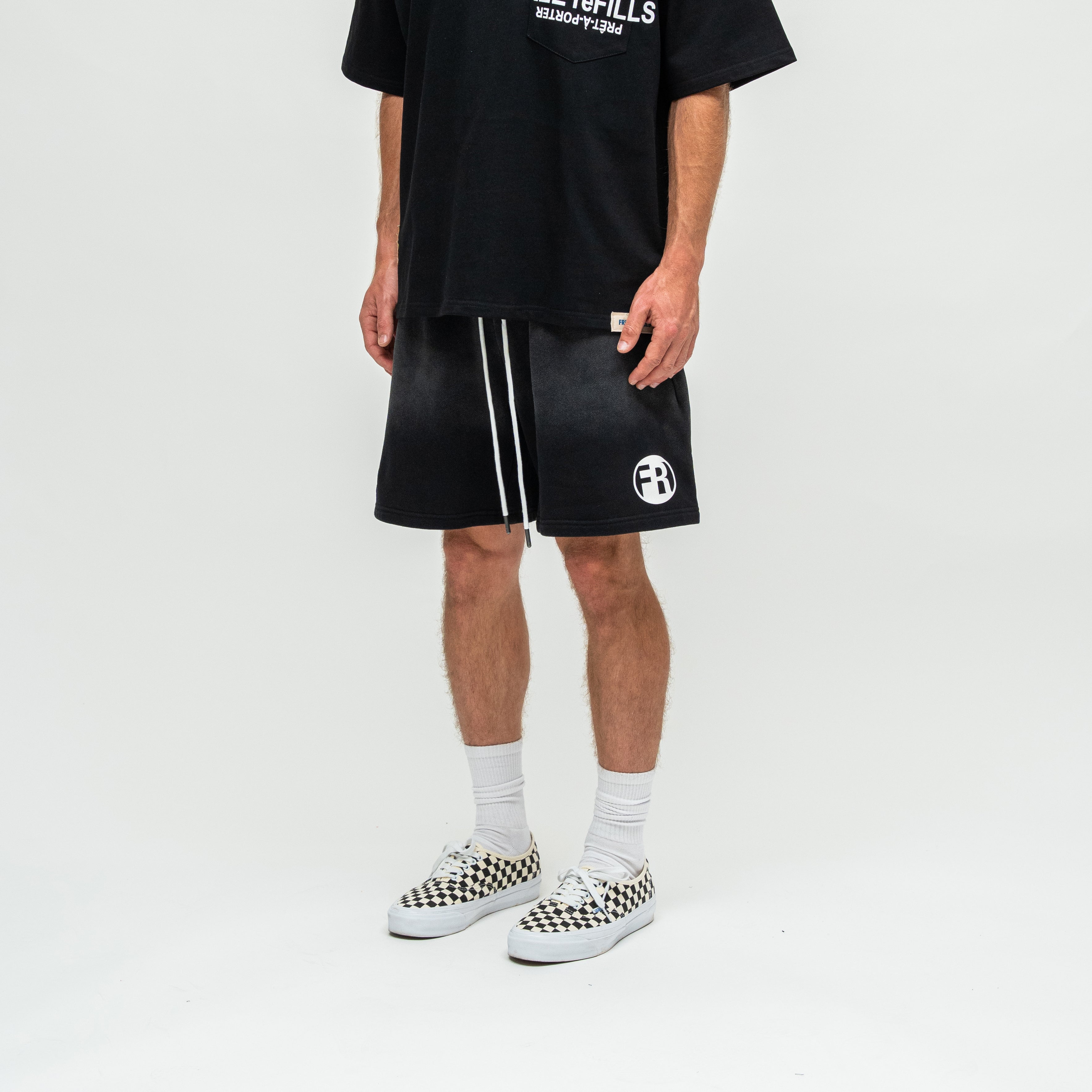 A model wearing black athletic shorts with white side stripes and a white circular logo, paired with a black graphic t-shirt, white socks, and black-and-white checkered Vans sneakers, shot against a plain white studio background.