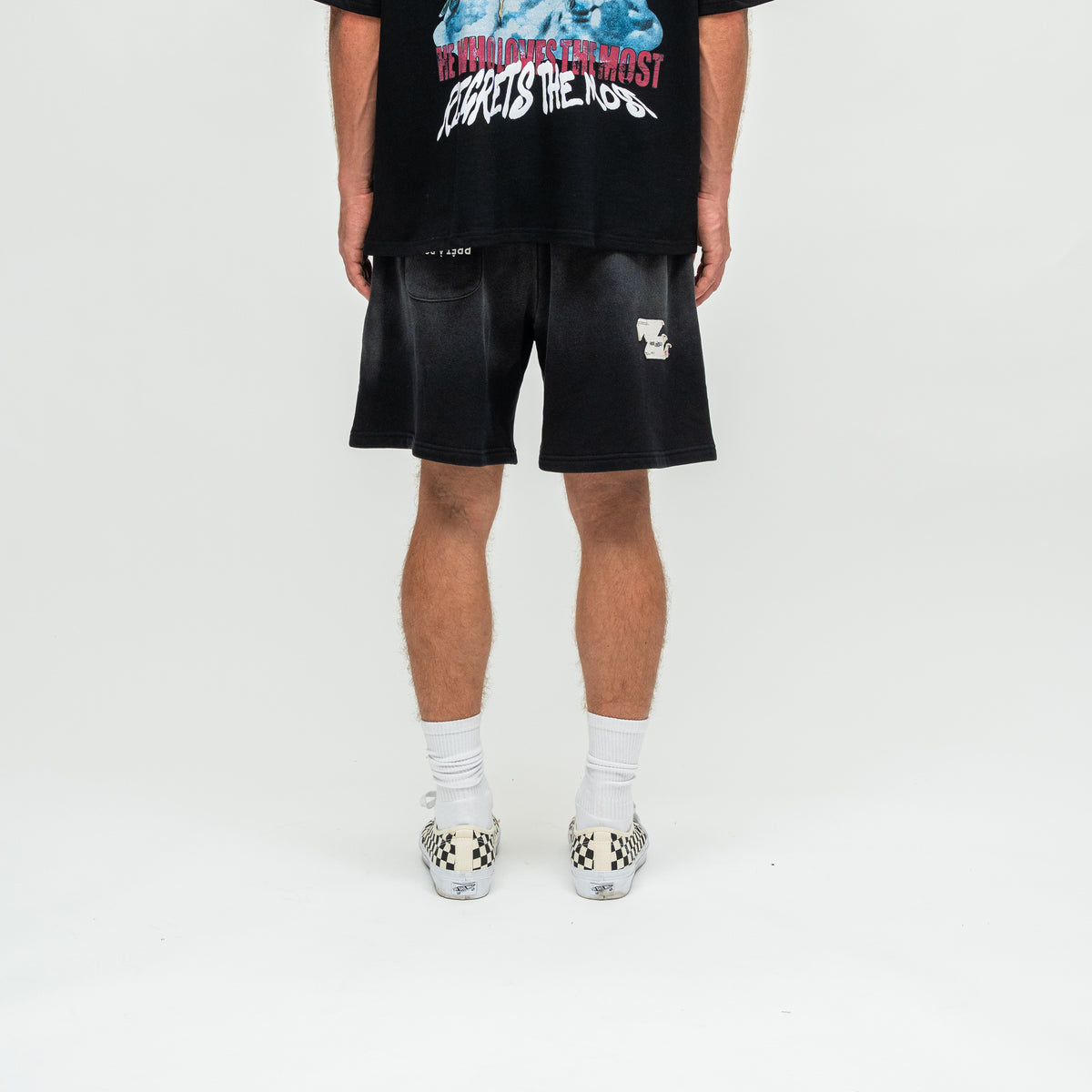A person shown from the waist down wearing black shorts with a faded wash, white crew socks, and checkerboard slip-on shoes, standing against a light gray studio background.