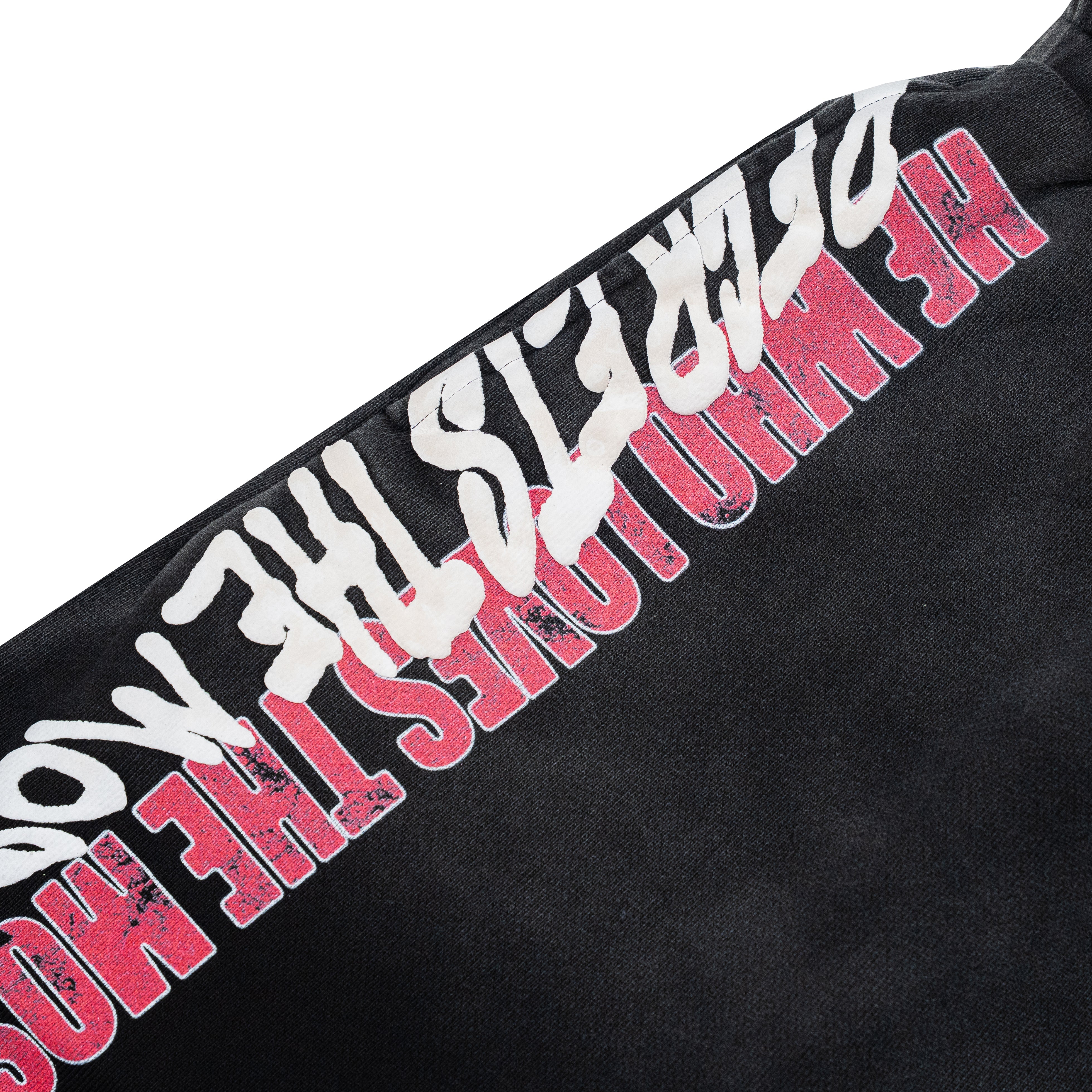 Close-up of a black garment featuring a diagonal graphic print with distressed white text and bold pink text reading 'HE WHO LOVES THE MOST.