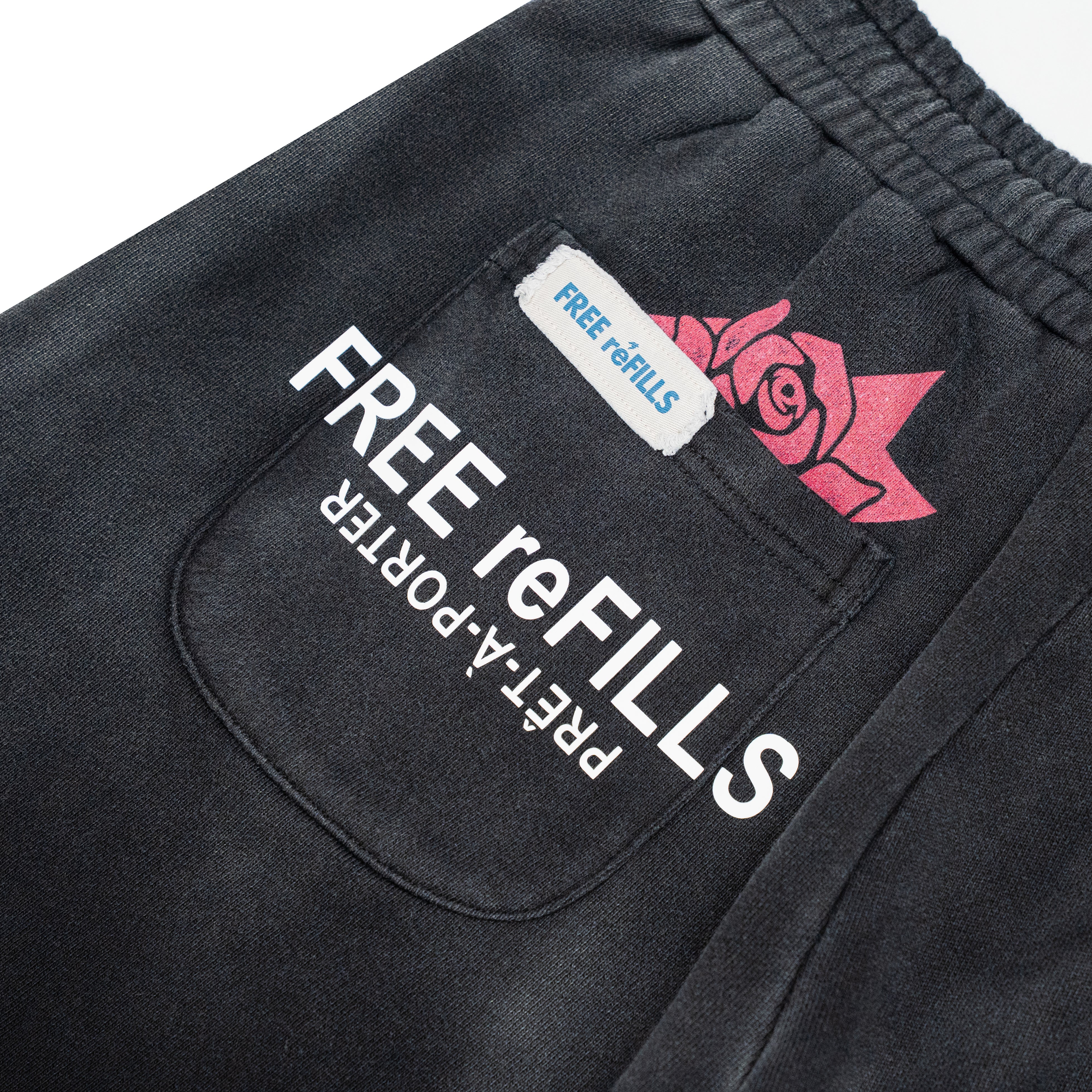 Close-up of a black sweatshirt pocket with white 'FREE reFILLS' text, a small white 'FREE reFILLS' label, and a pink rose graphic on the back pocket of black sweatpants.