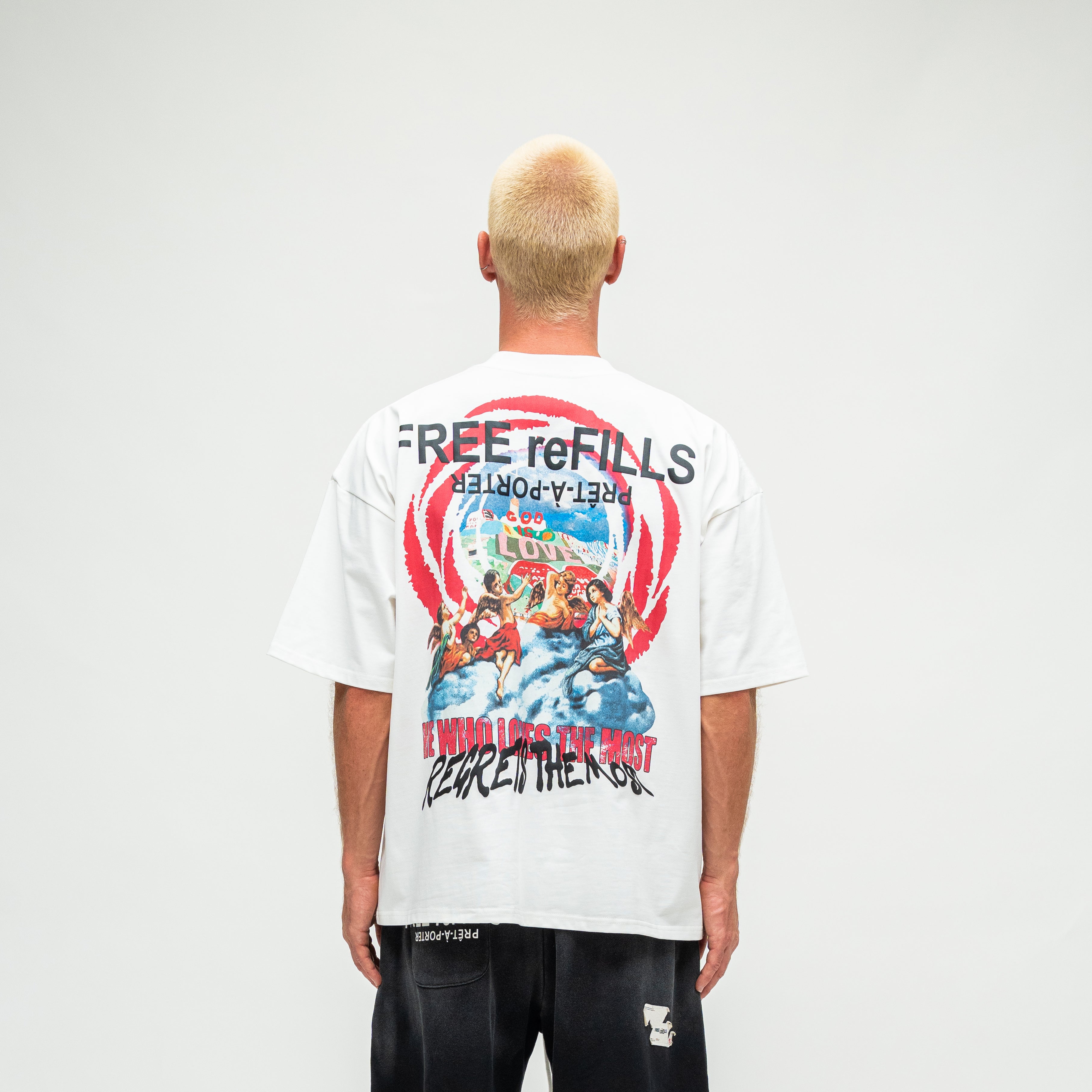 A man viewed from behind wearing a white graphic t-shirt with a large multicolor print on the back, paired with black shorts, against a plain light gray studio background.