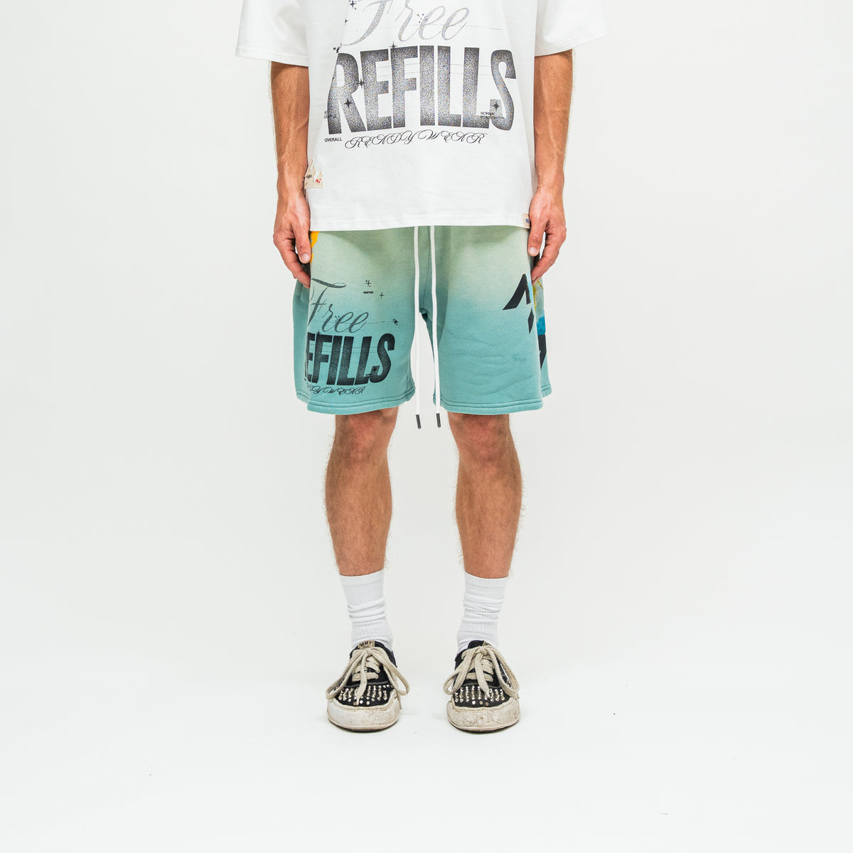 A person wearing teal and white gradient graphic shorts with 'Free REFILLS' text, a white graphic t-shirt, white socks, and black-and-white sneakers, standing against a plain white studio background.