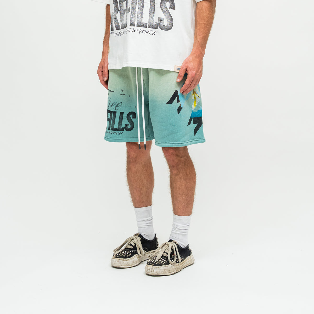 A person wearing Free Refills shorts with a teal-to-light-blue gradient and graphic print, styled with a white graphic t-shirt, white socks, and black-and-white sneakers against a white studio background.