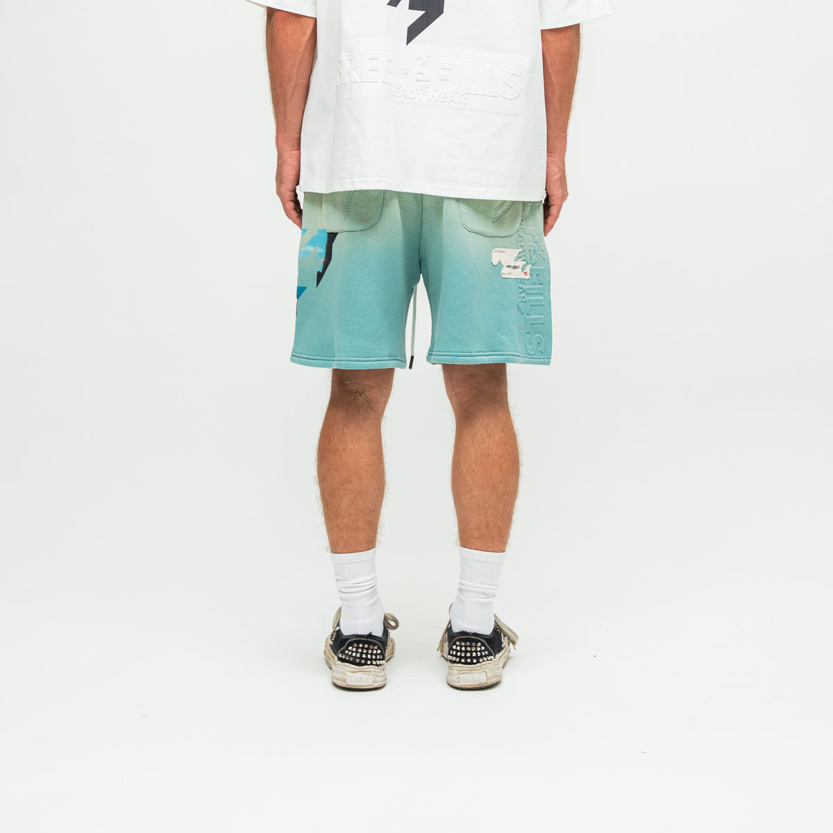 A person shown from the waist down wearing mint green and teal gradient shorts, a white graphic t-shirt, white socks, and black studded sneakers against a white studio background.