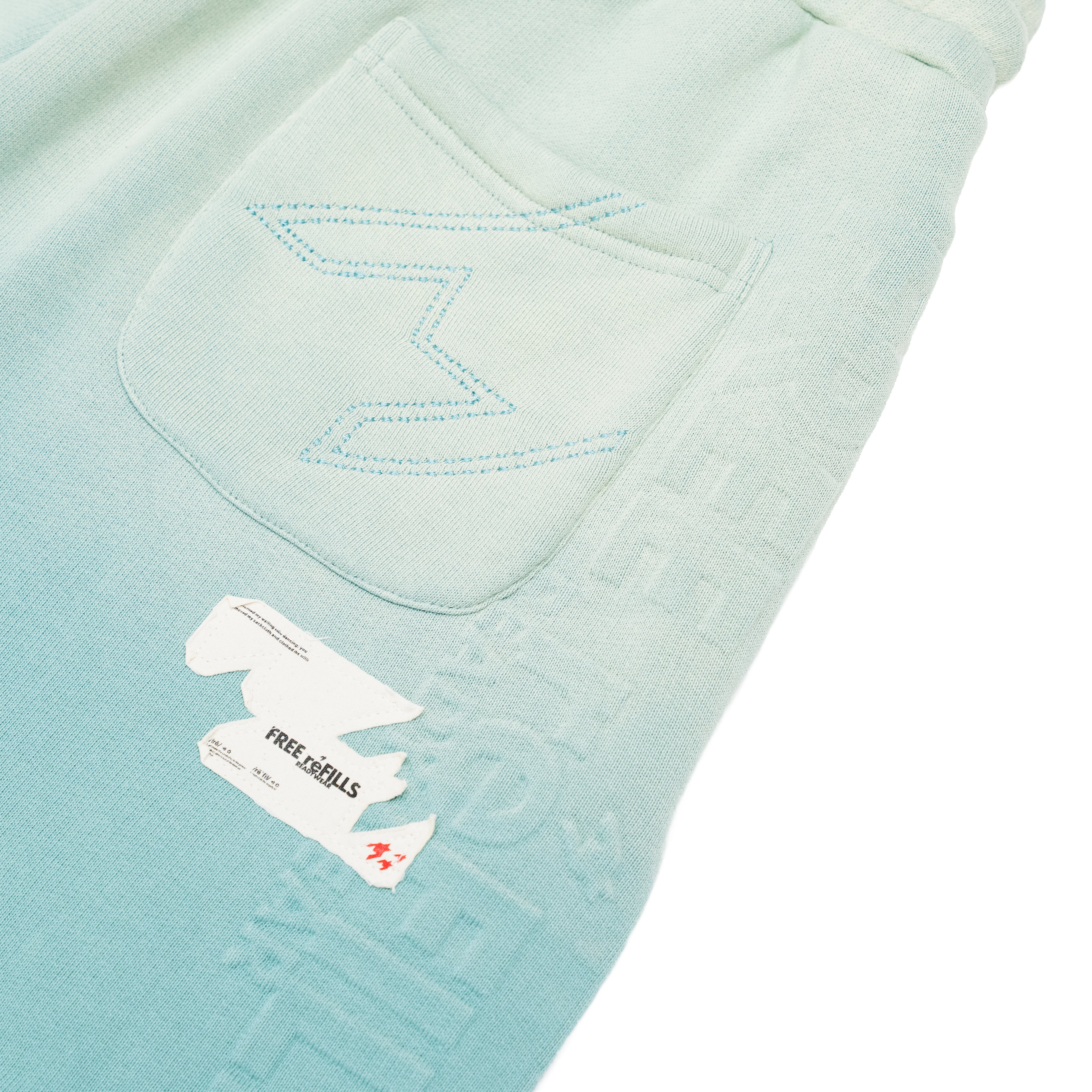 Close-up of a light mint green knit shorts, showing a pocket with decorative zigzag stitching and a white sewn-on brand tag that reads FREE REFILLS.
