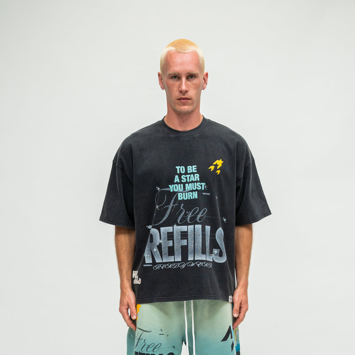 A man modeling a dark charcoal gray graphic t-shirt with 'Free REFILLS' and 'TO BE A STAR YOU MUST BURN' printed on the front, paired with light blue gradient shorts, against a plain light gray background.