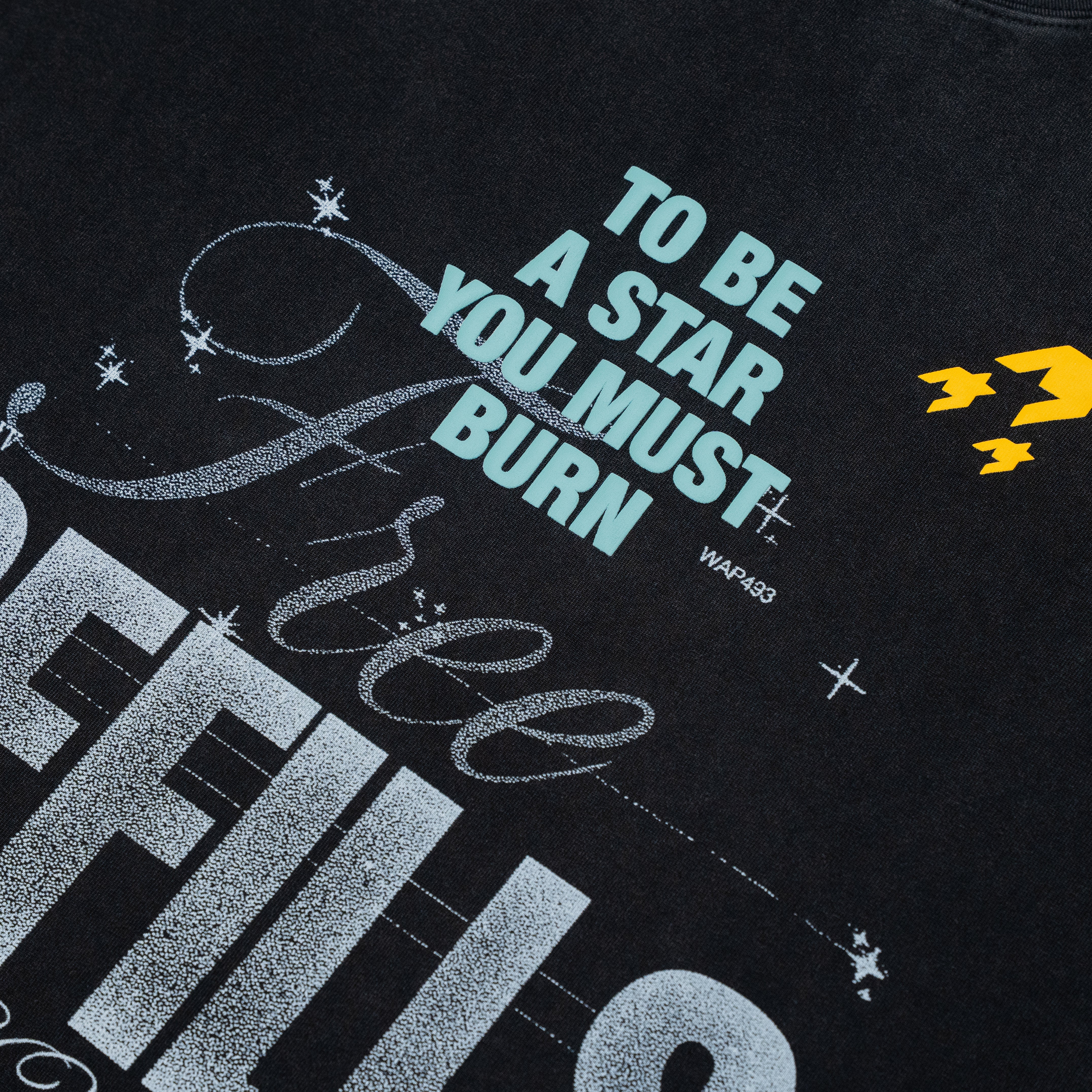 Close-up of a black graphic t-shirt featuring the text 'TO BE A STAR YOU MUST BURN' in light blue, distressed silver script, and a small yellow logo on the right side.